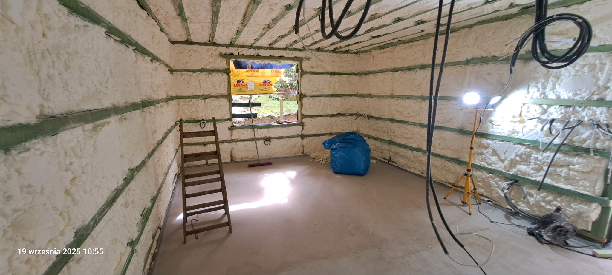 Wnętrze budynku w trakcie ocieplania pianką PUR. Widoczne drewniane ramy, okno z folią URSA, drabina, lampa na statywie i niebieski worek na odpady.