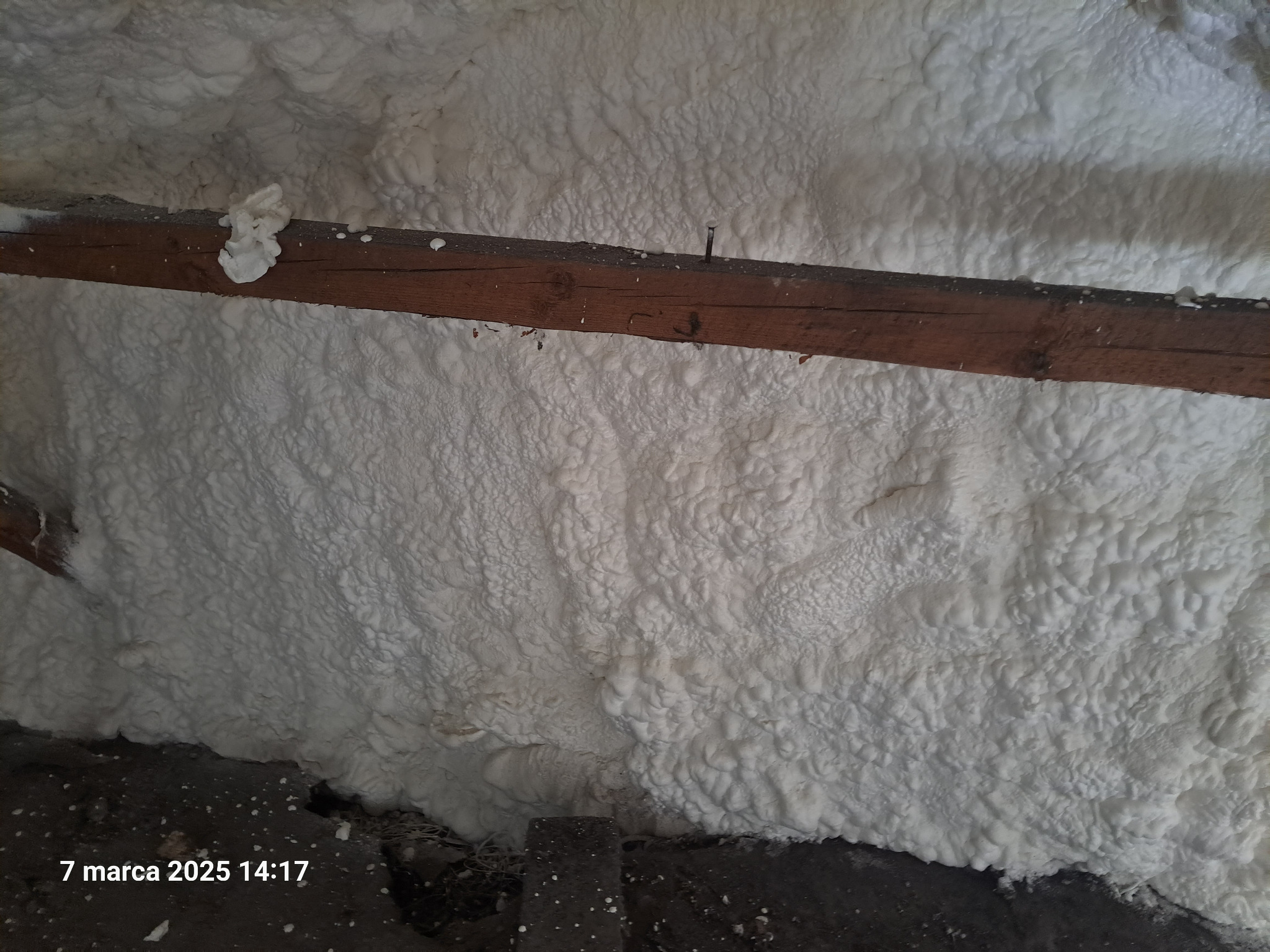 Szczegółowe ujęcie białej pianki izolacyjnej PUR na poddaszu, z widoczną drewnianą belką i wystającym gwoździem. Tekstura pianki przypomina gęstą, nierówną warstwę.