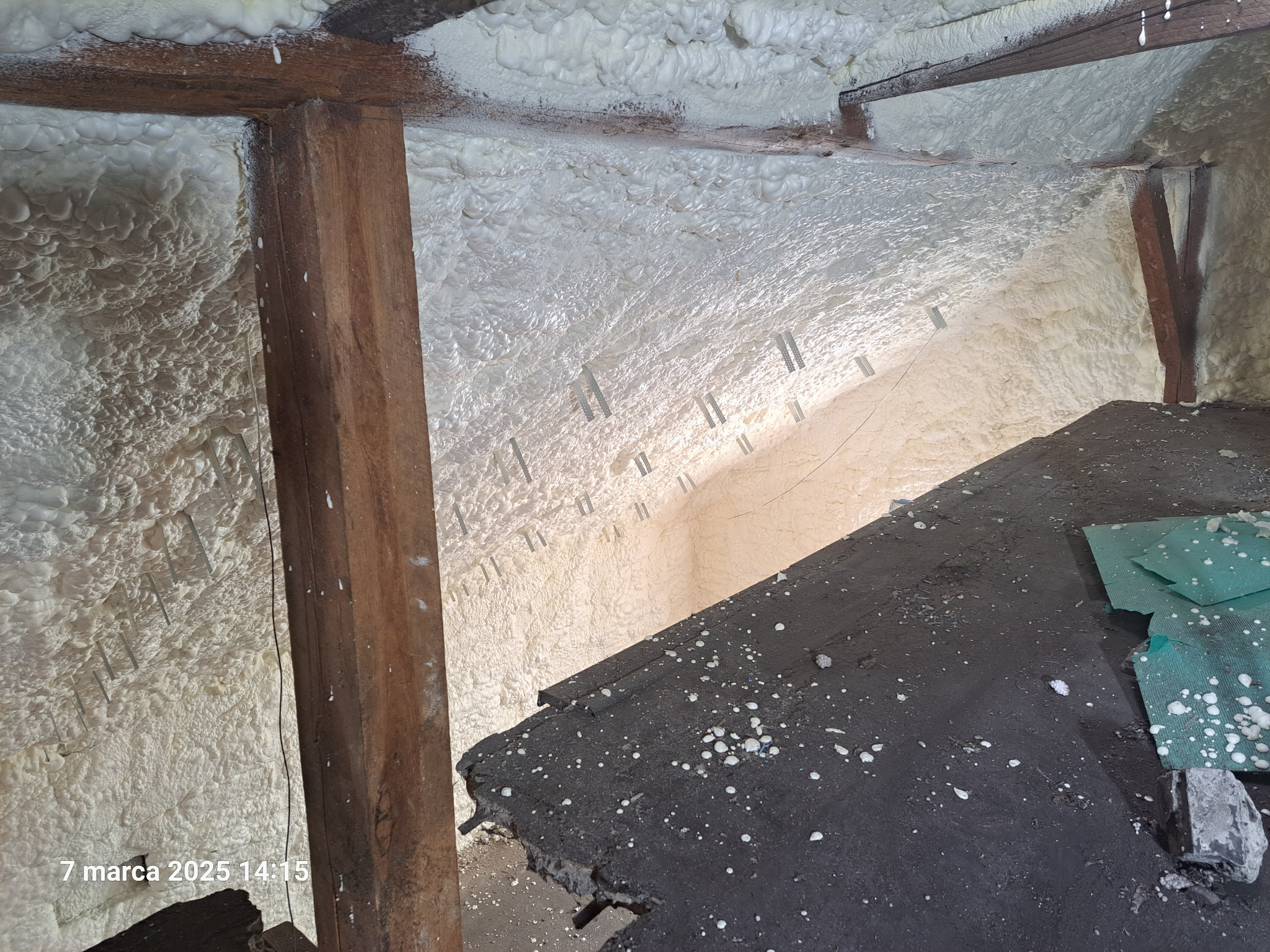 Ocieplanie poddasza pianką PUR z widocznymi elementami konstrukcji drewnianej i fragmentem podłogi. Izolacja natryskowa na skosach dachu.