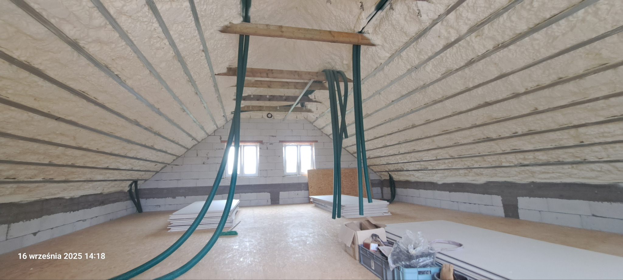 Poddasze po ociepleniu pianką PUR, widoczne rury do aplikacji, bloki betonowe i płyty kartonowo-gipsowe. Ocieplony dach z metalowymi profilami.