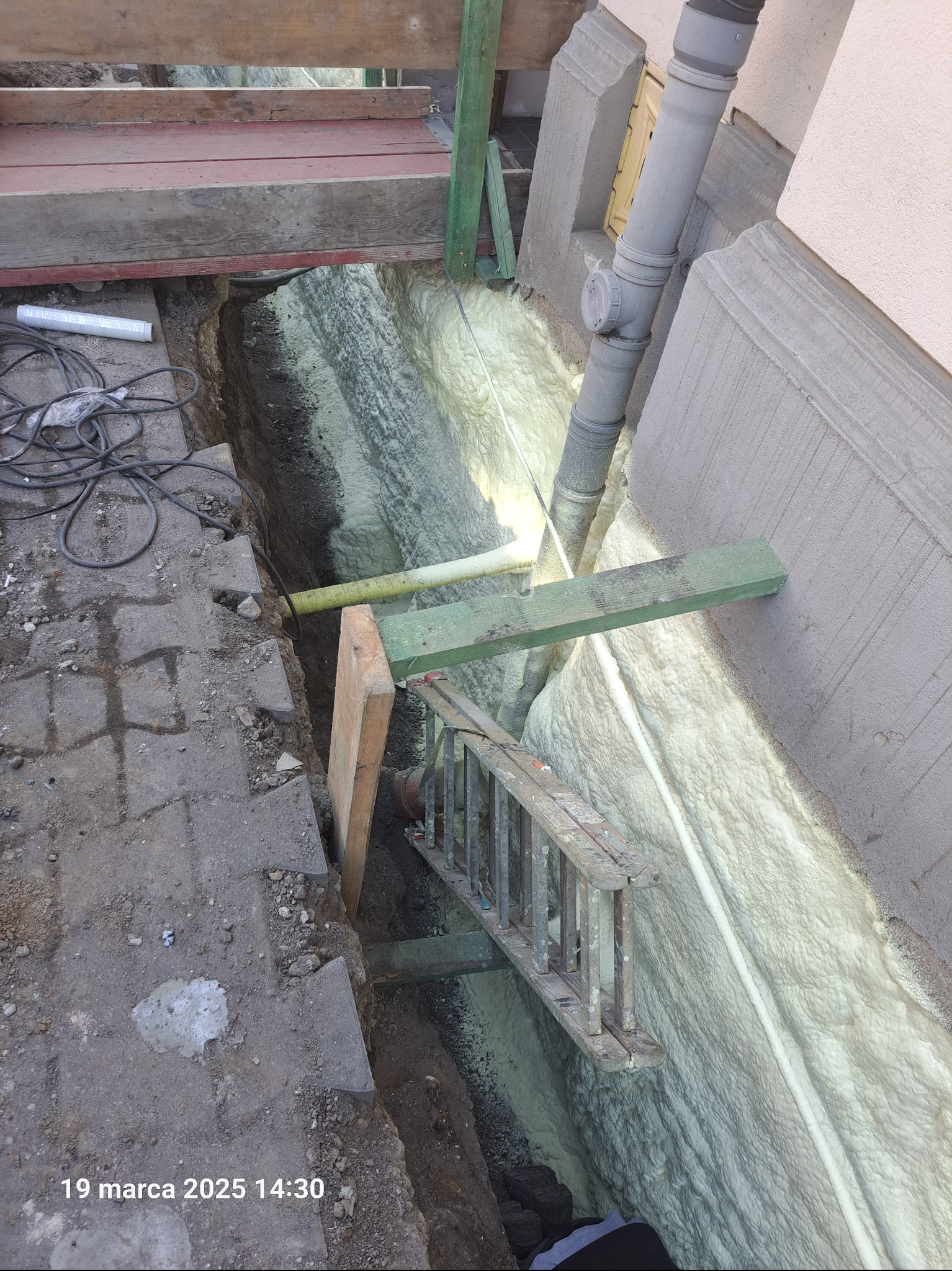 Fundament ocieplony pianką PUR, widoczny rów z izolacją, drabina i fragment ściany budynku. Detal: żółta rura wystająca spod elewacji.
