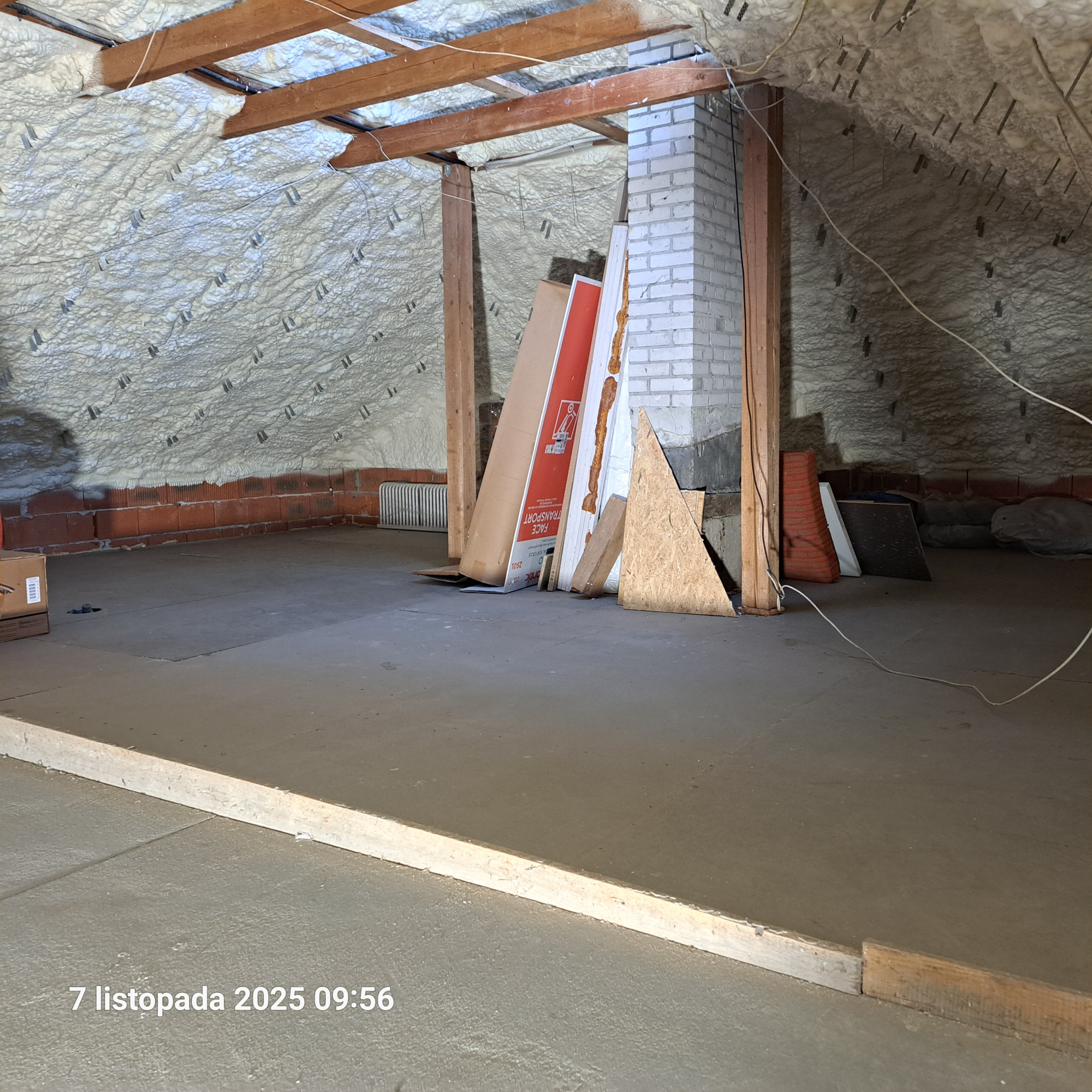 Poddasze po ociepleniu pianką PUR, widoczne drewniane elementy konstrukcyjne i biała pianka na skośnych ścianach. Na podłodze leżą płyty i inne materiały budowlane.