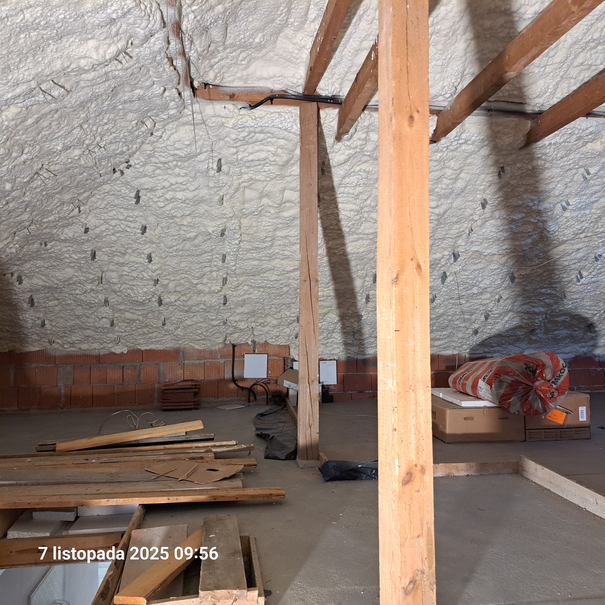 Poddasze po natrysku pianki PUR, widoczne drewniane elementy konstrukcyjne i fragment ściany z cegły. Biała, nierówna powierzchnia pianki pokrywa całą przestrzeń.