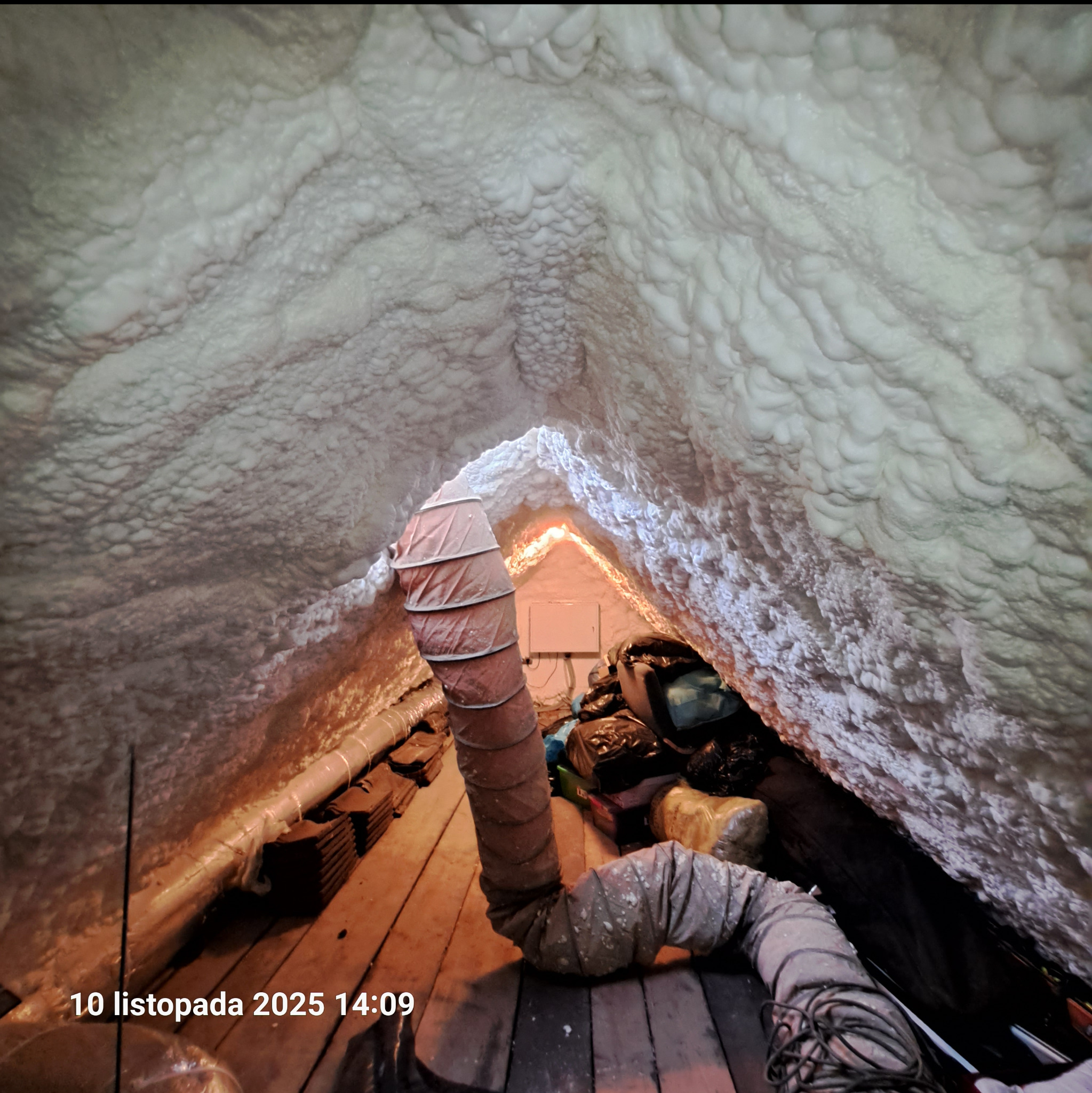 Poddasze po natrysku pianki PUR, widoczna struktura izolacji i przewód wentylacyjny. Oświetlenie podkreśla nierówności piany, tworząc unikalny efekt wizualny.