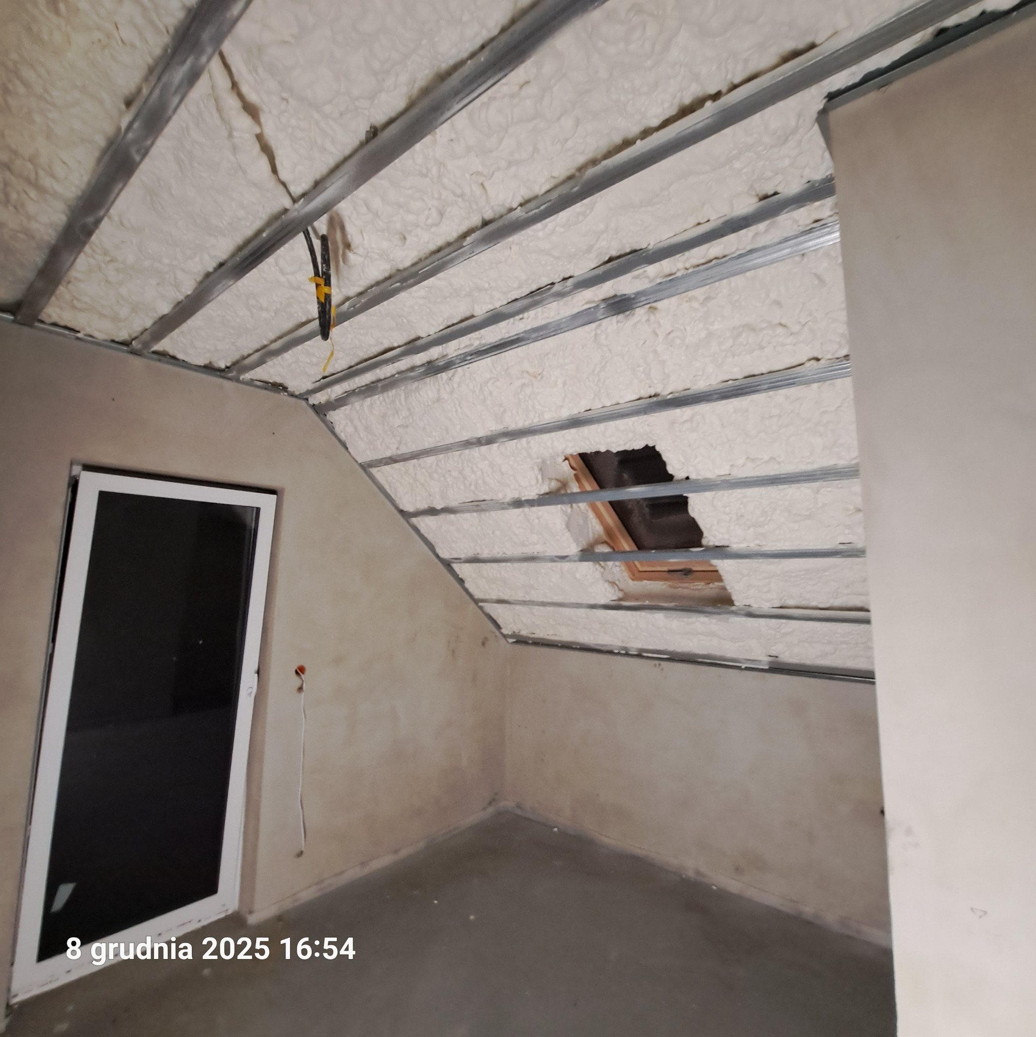 Poddasze z widoczną izolacją pianką PUR między metalowymi profilami, okno dachowe z drewnianą ramą. Widoczne fragmenty ścian i okno w białej ramie.