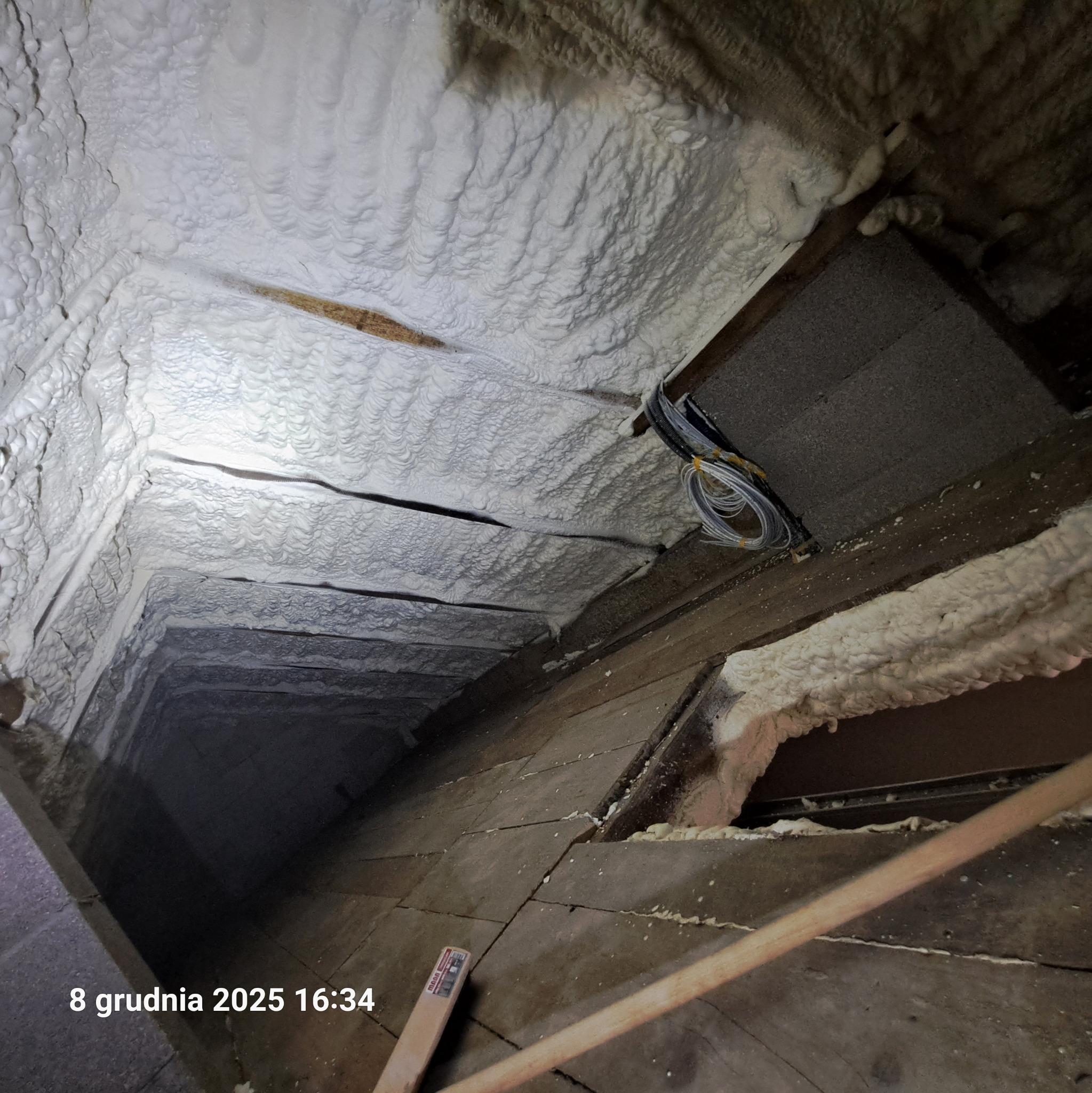 Strych po natrysku pianki PUR, widoczne drewniane elementy konstrukcyjne i przewody. Biała, nierówna powierzchnia izolacji. Ujęcie z dołu, perspektywa.