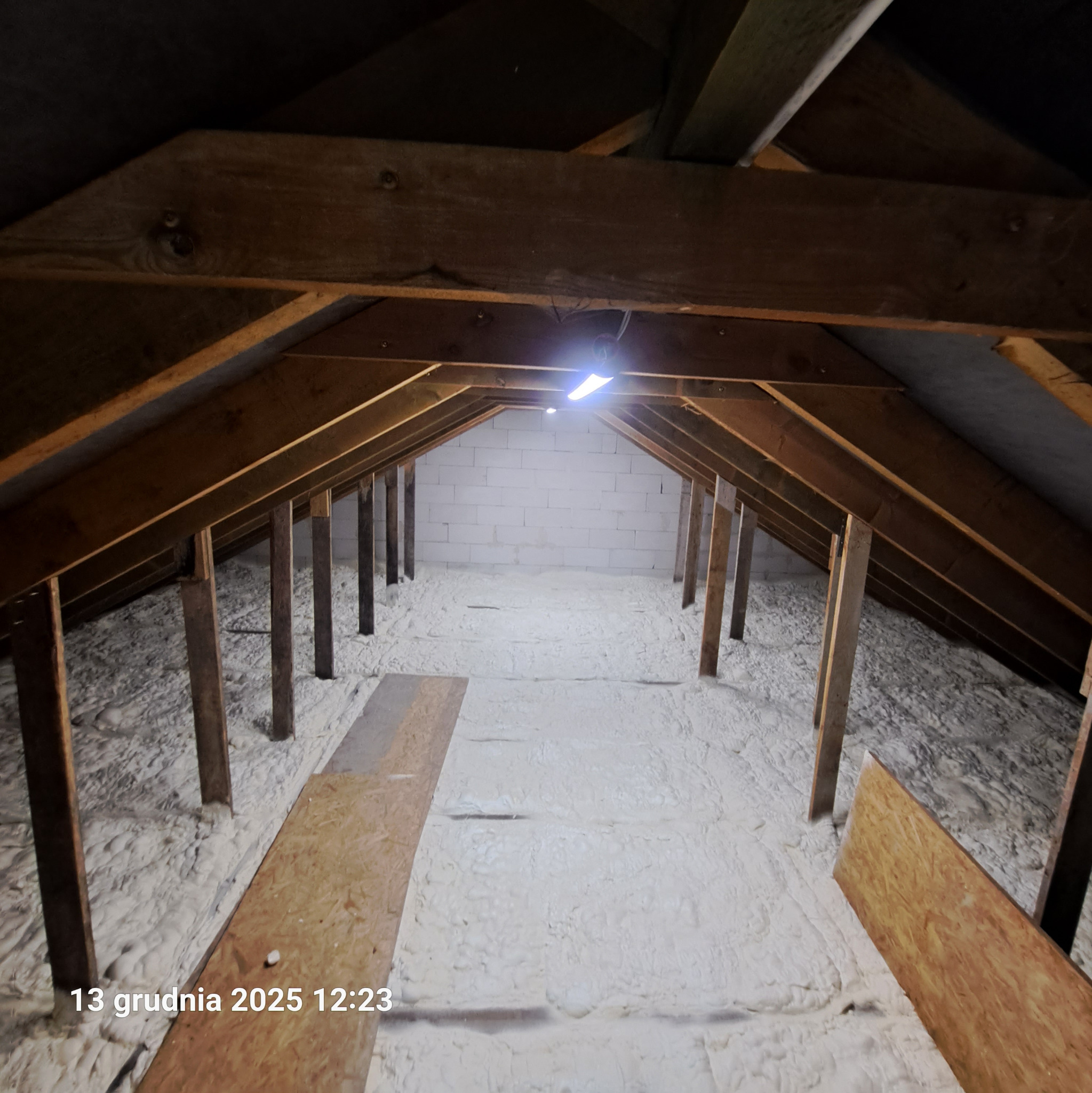 Poddasze z drewnianą konstrukcją i białą pianką izolacyjną, widok z góry. Widoczne deski na podłodze i oświetlenie. Ściana z białych pustaków w tle.