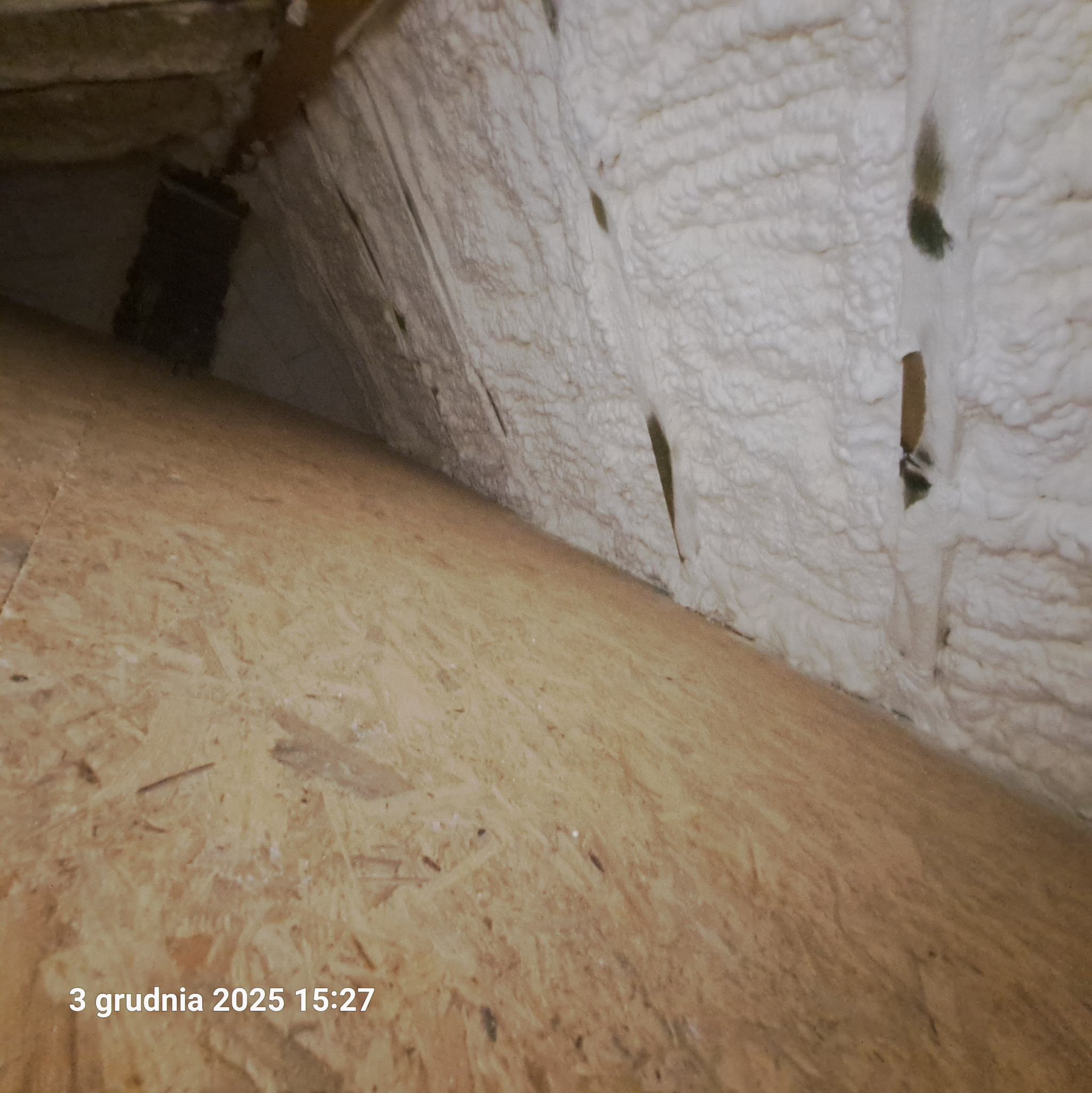 Skośny strych z deskami i białą pianką izolacyjną na ścianach, widoczne ciemne plamy. Ujęcie z dołu, data na zdjęciu: 3 grudnia 2025.