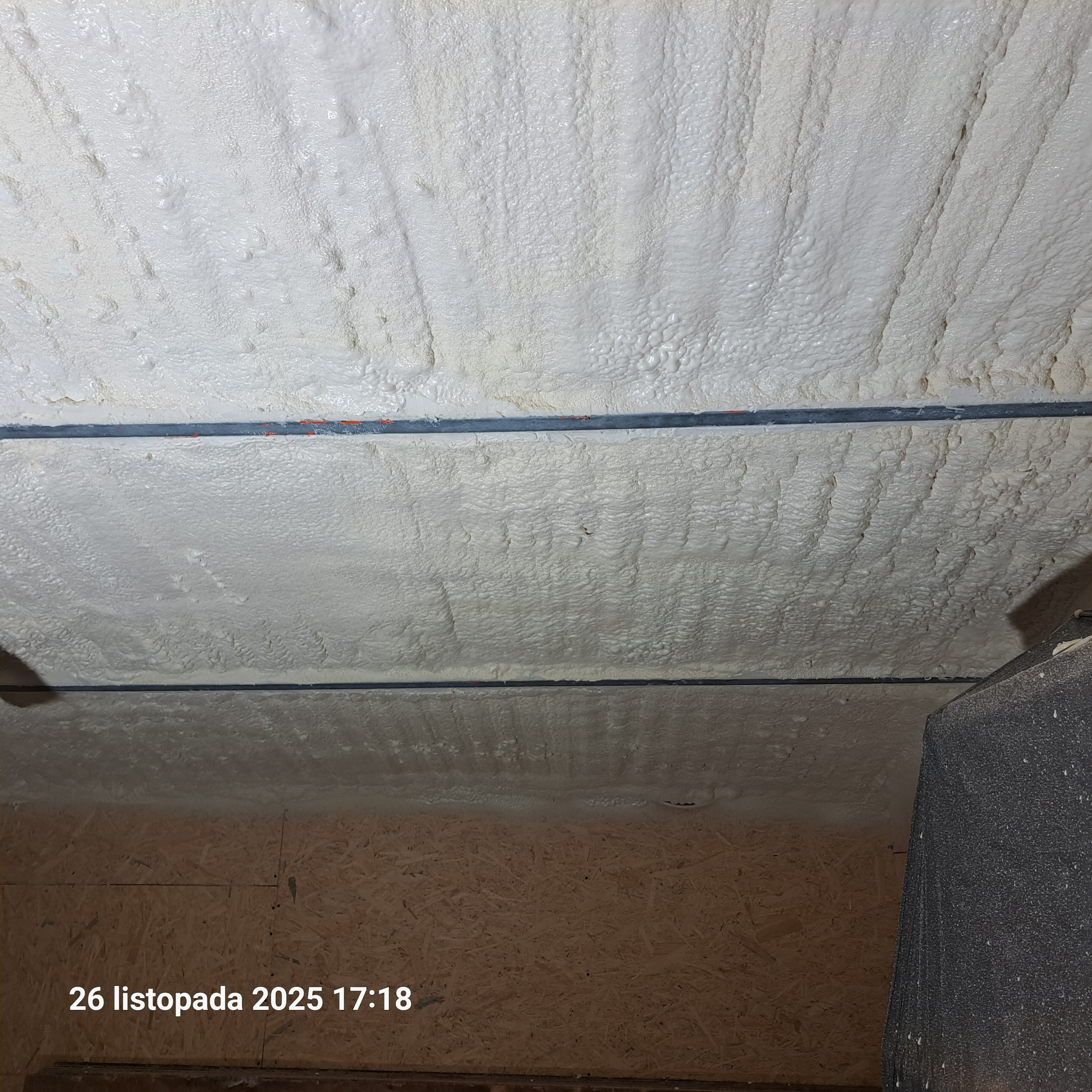 Szczegółowe ujęcie białej pianki izolacyjnej PUR na poddaszu, z widoczną strukturą i elementami konstrukcyjnymi. Widoczne ślady po natrysku pianki.