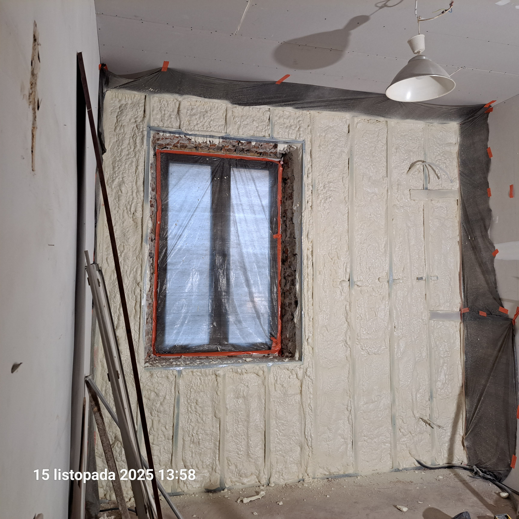 Ocieplanie pianką PUR ściany wewnętrznej z oknem, widoczne profile i folia ochronna. Świeżo nałożona pianka o jasnej barwie, sufit z płyt kartonowo-gipsowych.
