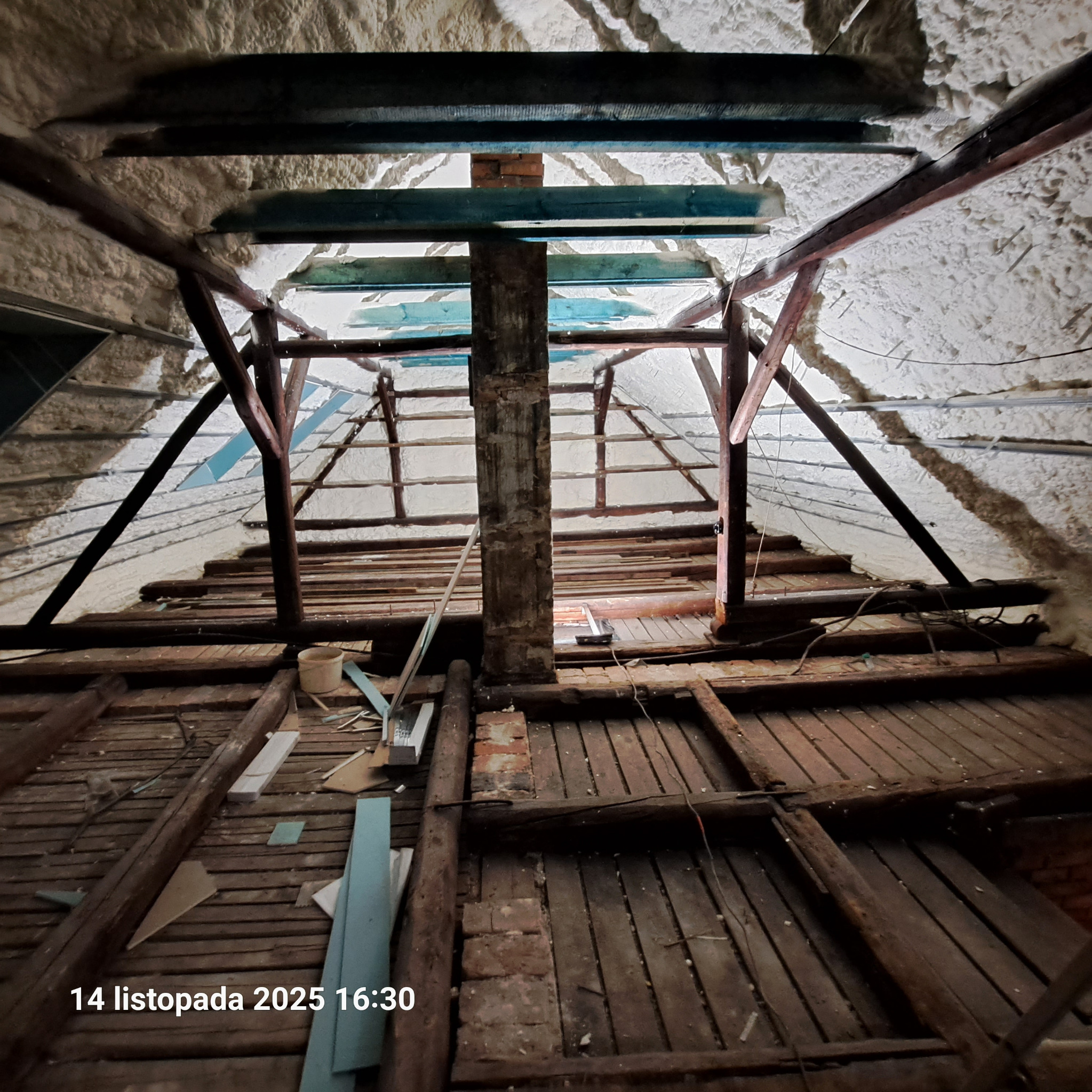 Poddasze z drewnianą konstrukcją dachu, częściowo ocieplone pianką PUR. Widoczne elementy starej konstrukcji i fragmenty izolacji. Ujęcie z dołu, perspektywa zbieżna.