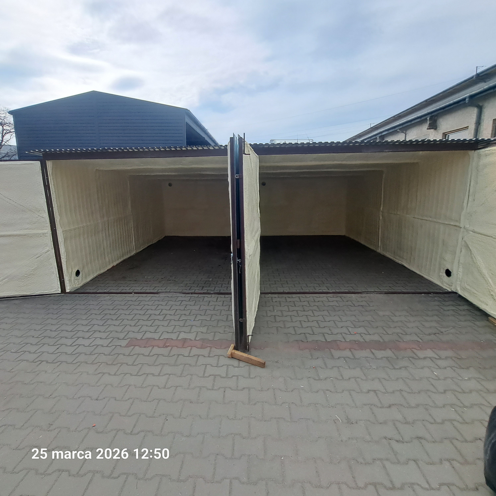 Otwarty garaż dwustanowiskowy po ociepleniu pianką PUR, widoczne ściany i brama pokryte jasną pianą. Na podłodze kostka brukowa, w tle budynki.