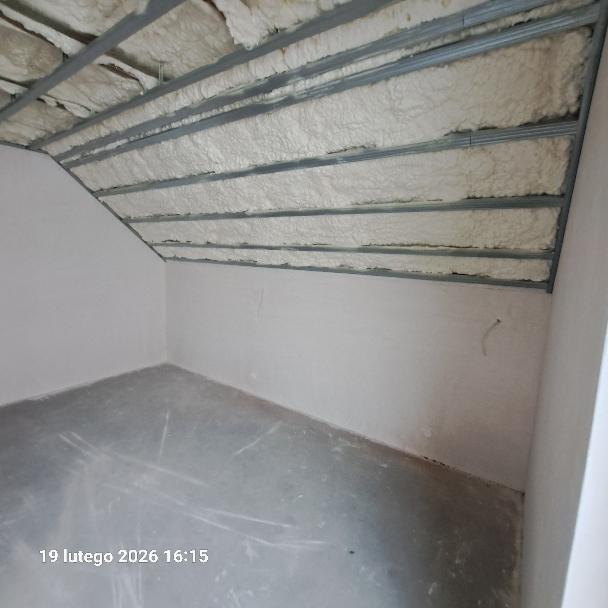 Poddasze z widoczną izolacją pianką PUR między metalowymi profilami. Ściany w kolorze złamanej bieli, surowa podłoga. Ujęcie z dołu, widoczny fragment daty.