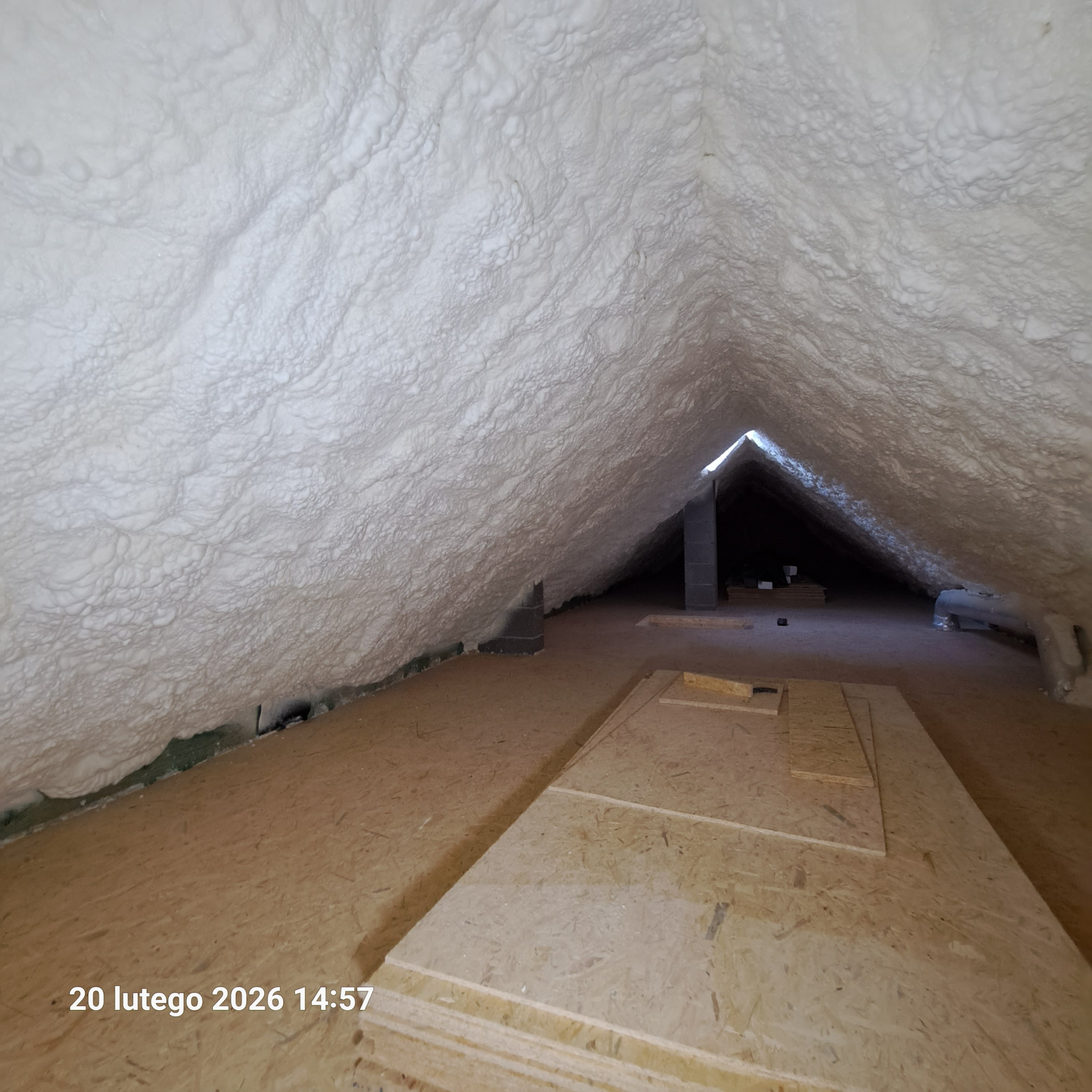 Poddasze po ociepleniu pianką PUR, widoczne płyty OSB na podłodze i szara ściana komina. Biała, nierówna struktura pianki pokrywa skośne ściany i sufit.