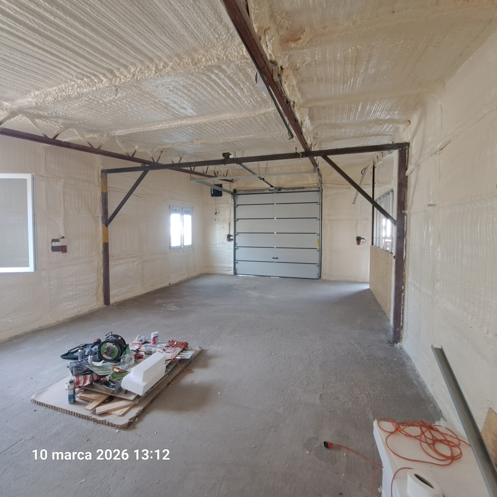 Wnętrze garażu po ociepleniu pianką PUR: ściany i sufit pokryte grubą warstwą izolacji, widoczne narzędzia na podłodze, brama garażowa zamknięta.