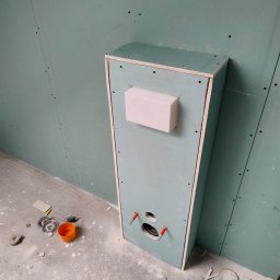 Marcin Gołąbek - Narożnik z zabudową z płyty karton-gips, widoczne otwory na instalacje. Ściany wykończone płytami gipsowymi, podłoga surowa, narzędzia w pobliżu.