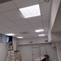 Marcin Gołąbek - Pracownik na drabinie montuje kaseton sufitowy w pomieszczeniu biurowym. Widoczne rusztowania, narzędzia i sufit podwieszany z oświetleniem.