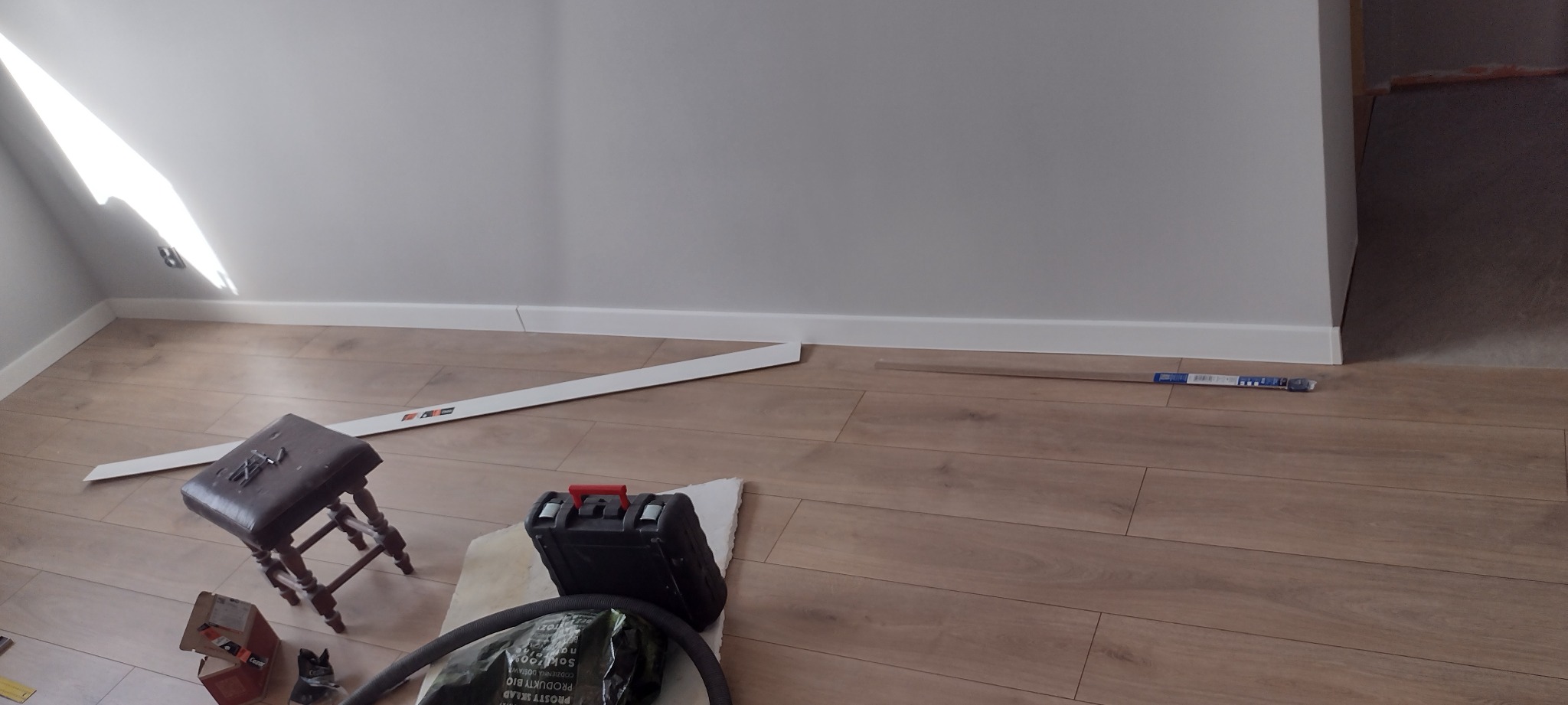 Fragment pokoju z ułożonymi panelami podłogowymi, białą listwą przypodłogową, narzędziami pracy, stołkiem z gwoździami i odkurzaczem w rogu pomieszczenia.