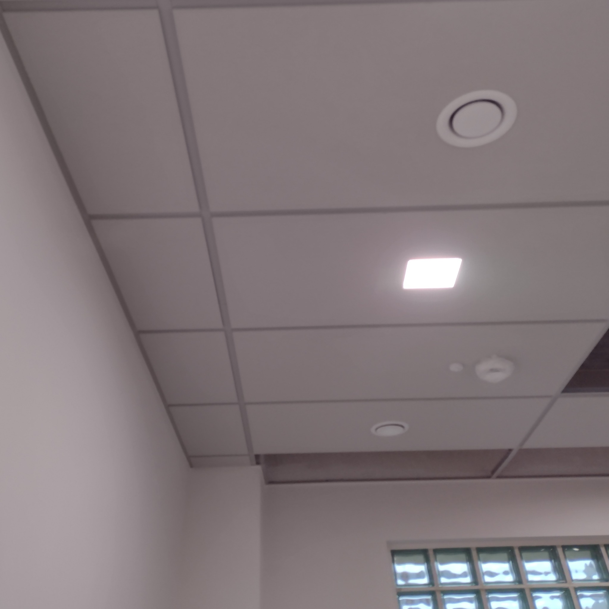 Kasetonowy sufit podwieszany z widocznymi punktami świetlnymi i kratką wentylacyjną w tle, z fragmentem ściany i okna z luksferami.