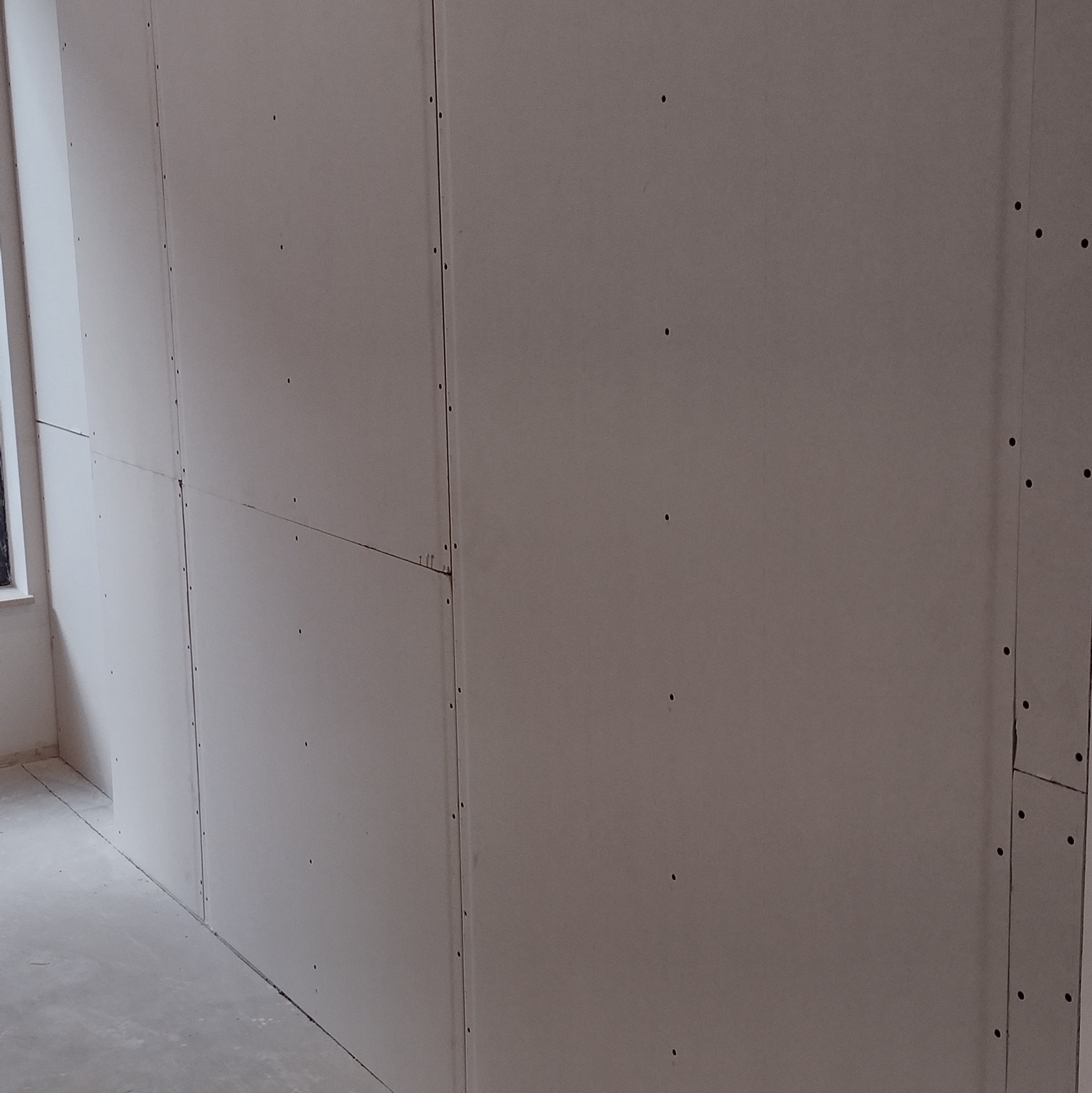 Ściana wewnętrzna obłożona płytami gipsowo-kartonowymi, widoczne wkręty mocujące oraz łączenia płyt, surowy stan przed dalszymi pracami wykończeniowymi.