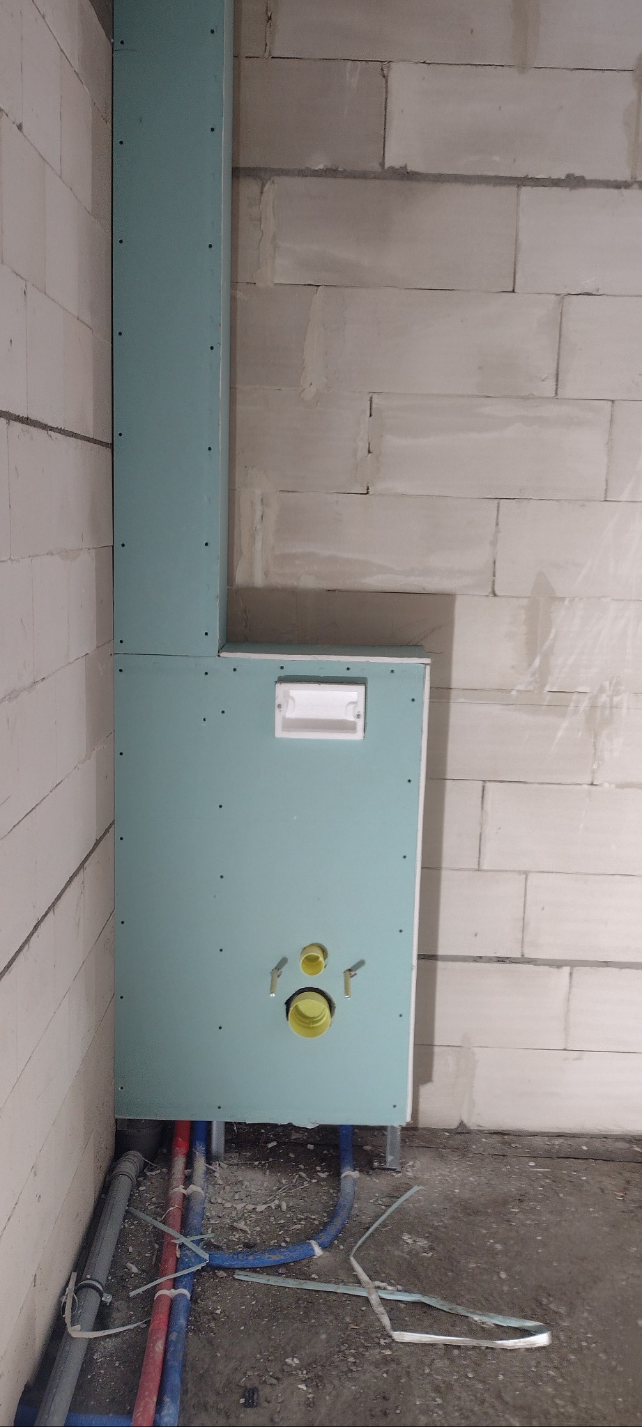 Niebieska płyta kartonowo-gipsowa zabudowująca stelaż podtynkowy WC, widoczne rury instalacyjne i otwory na przyłącza, ściana z bloczków betonowych w tle.