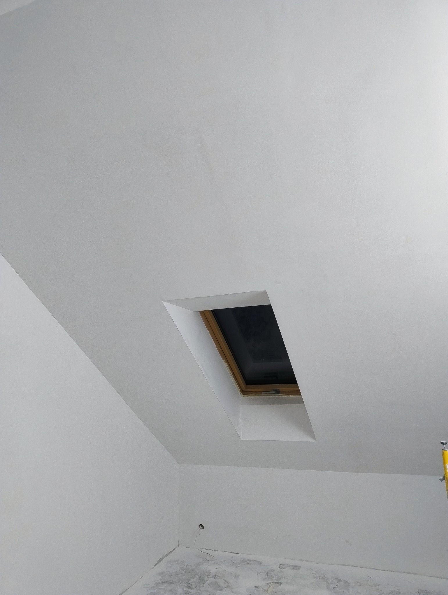 Poddasze po remoncie z oknem dachowym, ściany i sufit pokryte świeżą, białą warstwą, widoczne ślady po pracach na podłodze, fragment drabiny po prawej.