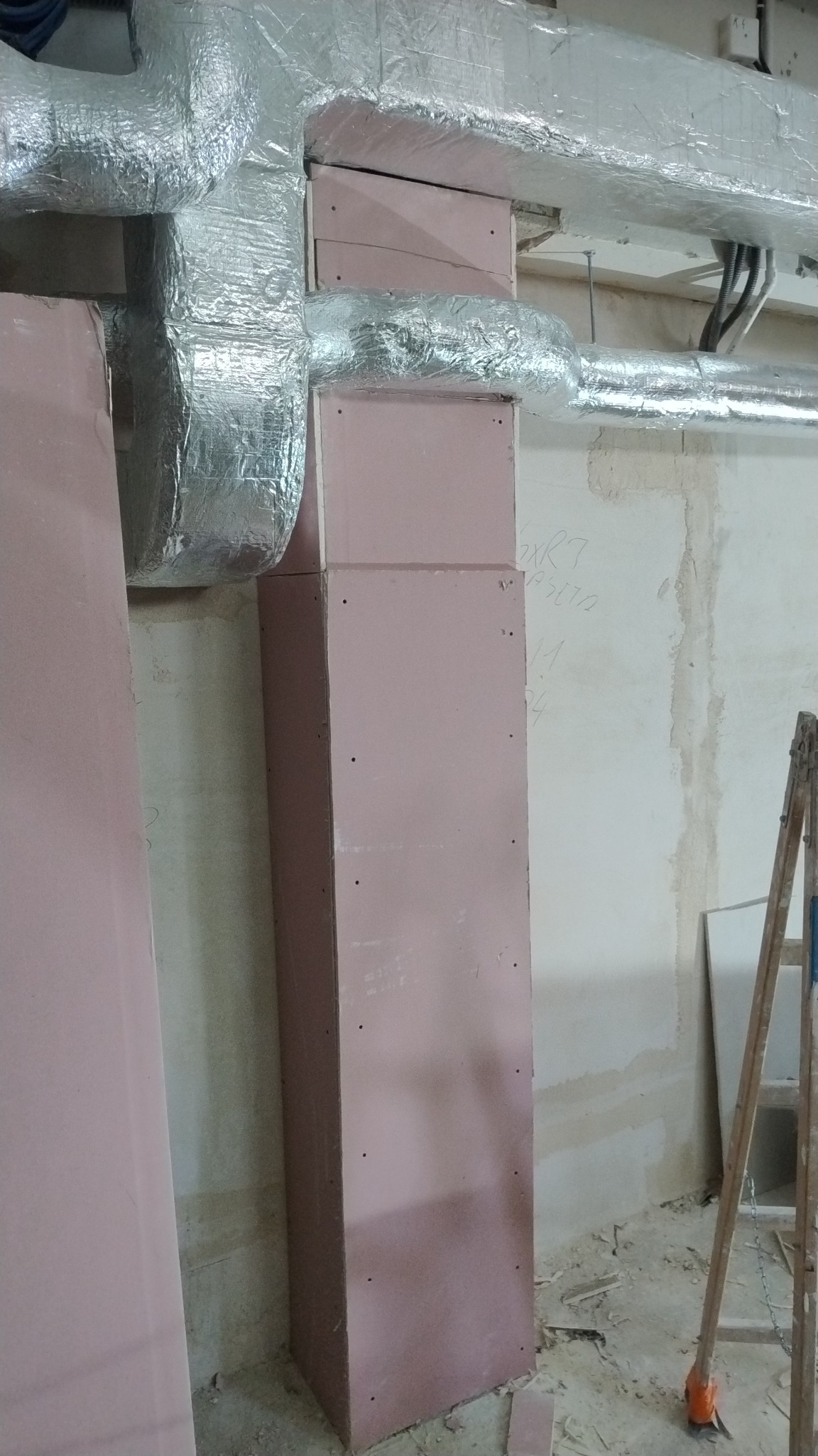 Obudowa z płyt gipsowo-kartonowych maskująca instalację wentylacyjną, widoczne srebrne rury i drabina oparta o ścianę w surowym wnętrzu.