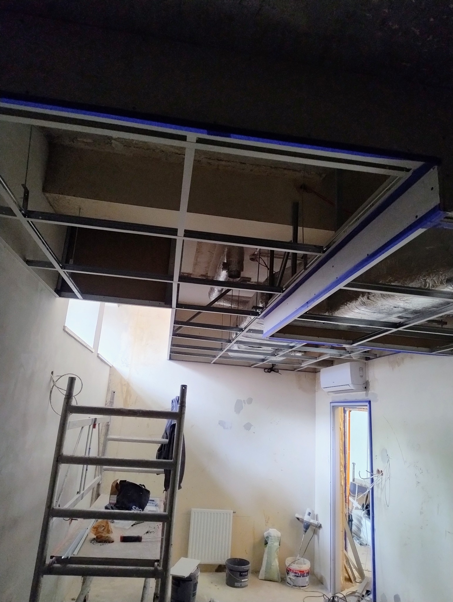 Montaż sufitu podwieszanego z widoczną konstrukcją metalową i instalacjami. U dołu drabina i narzędzia. Prace wykończeniowe w pomieszczeniu.
