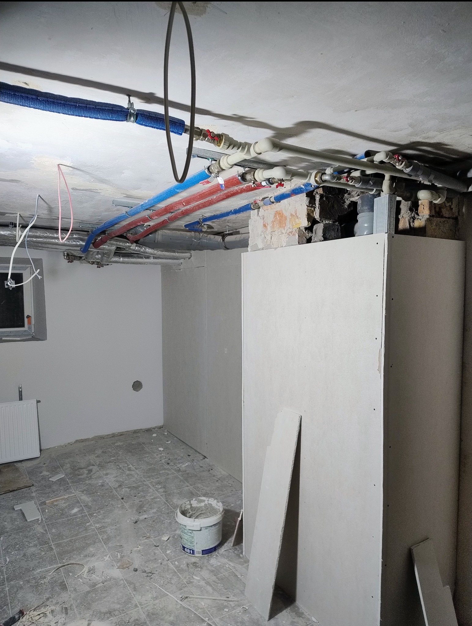 Wnętrze w trakcie remontu: widoczne instalacje pod sufitem, ściany przygotowane pod gładzie, fragment obudowy z płyty karton-gipsu i wiadro z zaprawą.