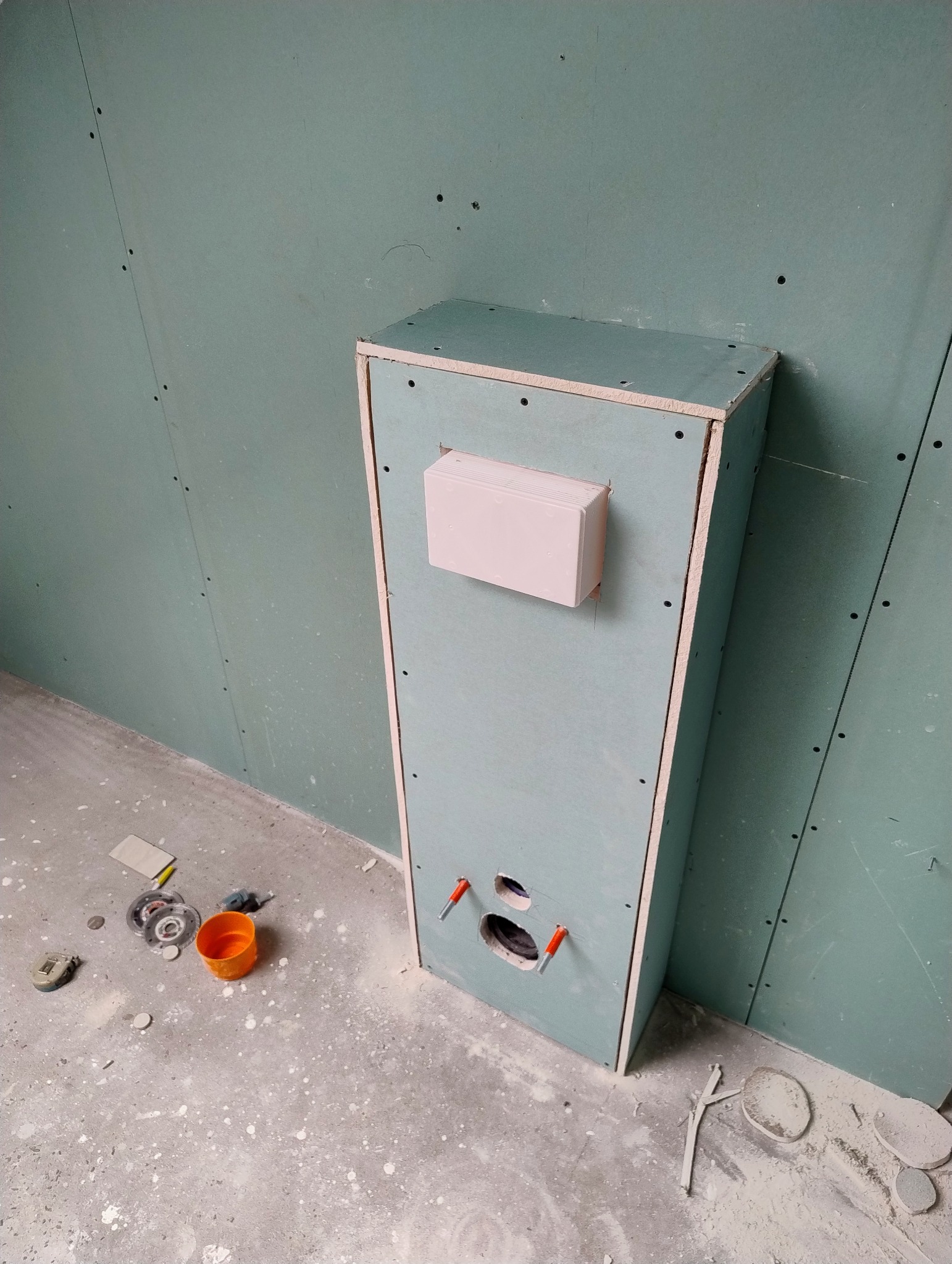 Narożnik z zabudową z płyty karton-gips, widoczne otwory na instalacje. Ściany wykończone płytami gipsowymi, podłoga surowa, narzędzia w pobliżu.