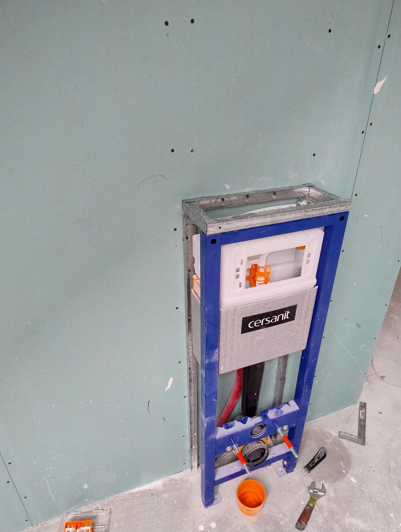 Stelaż podtynkowy Cersanit zamontowany przy ścianie z płyty karton gips, widoczne narzędzia i fragment podłogi. Instalacja przed zabudową.