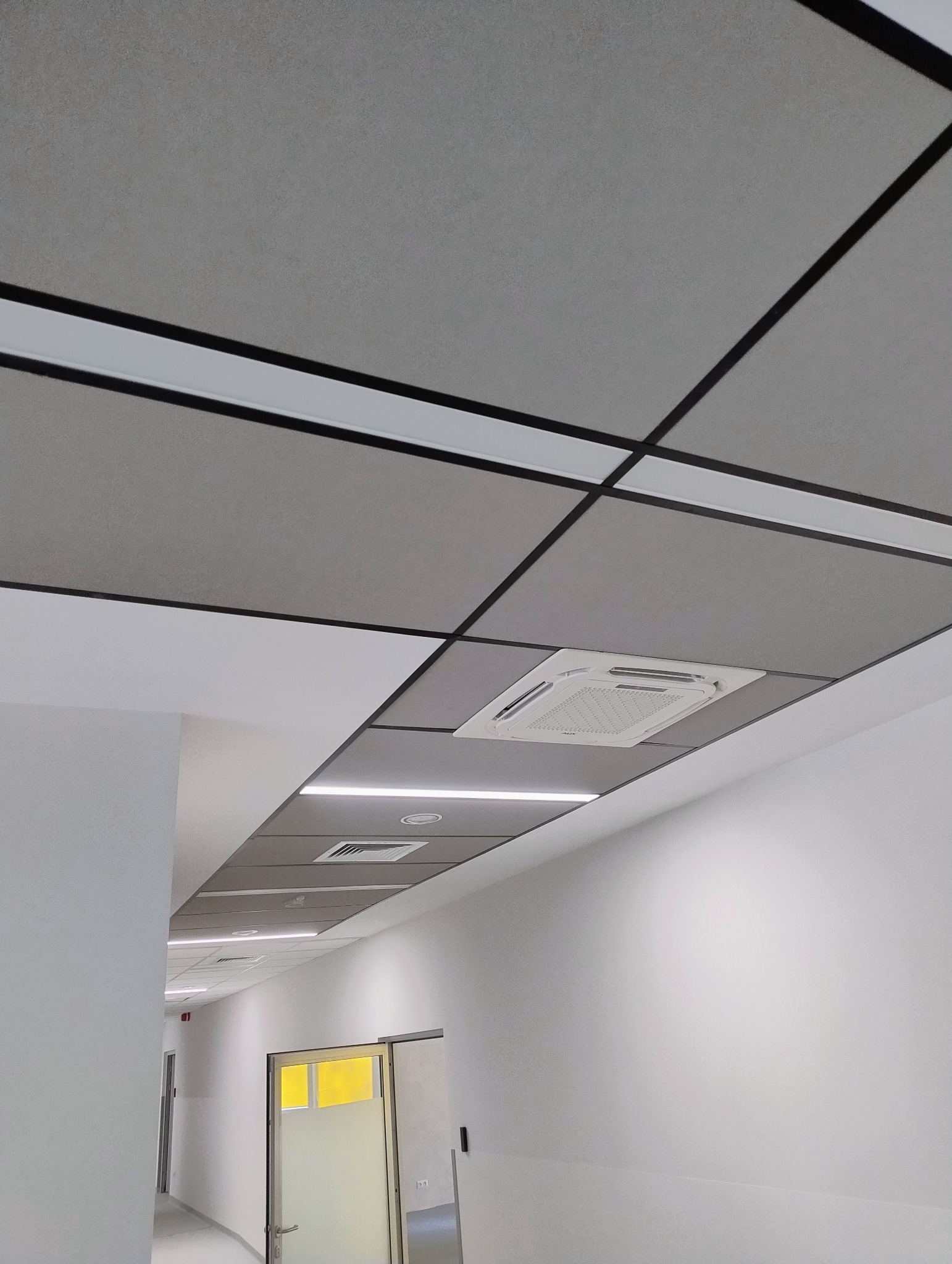 Sufit podwieszany z szarymi panelami i czarnymi listwami, oświetlenie LED i klimatyzacja w biurze. Perspektywa z dołu, widoczny fragment korytarza.