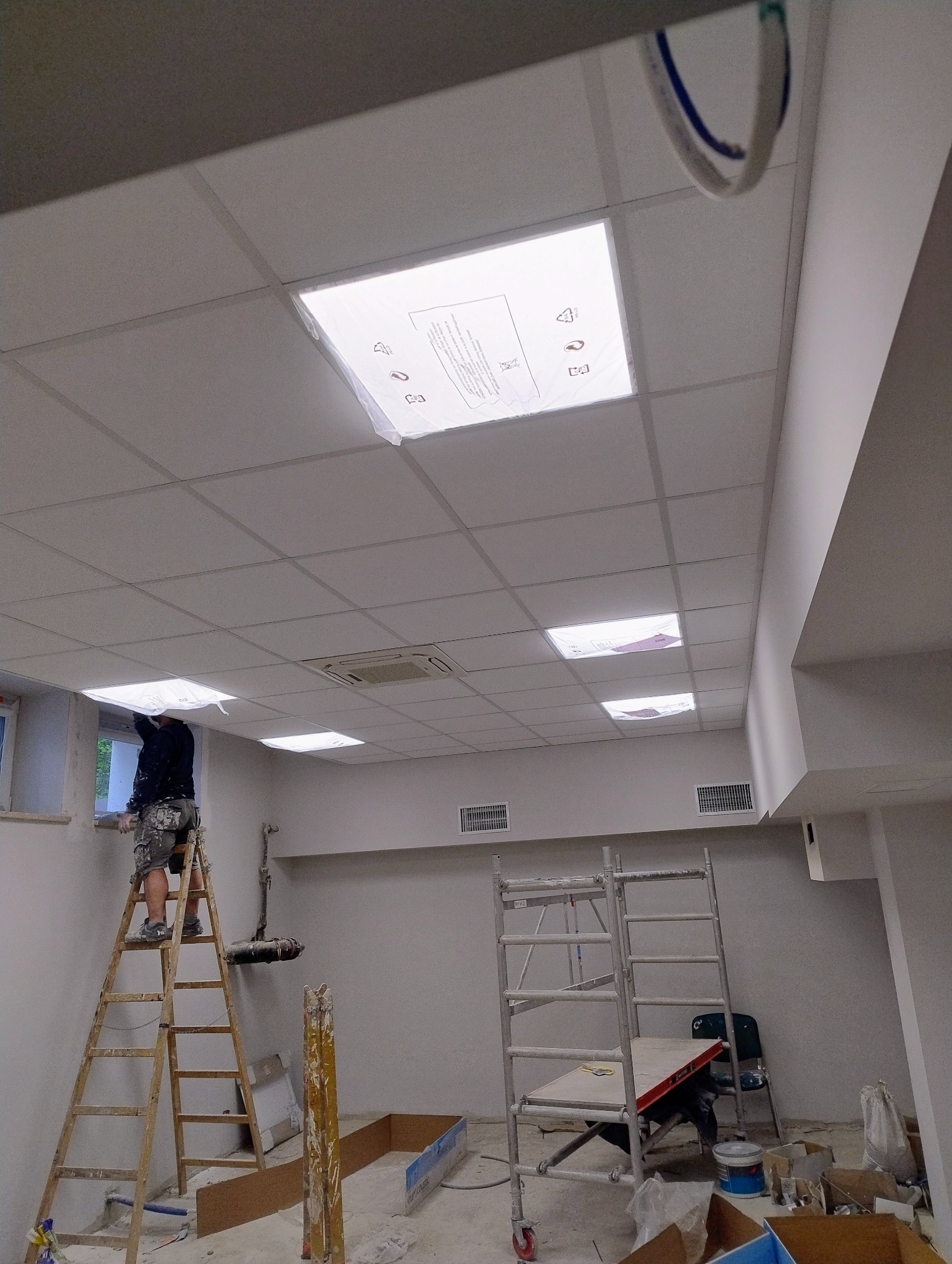 Pracownik na drabinie montuje kaseton sufitowy w pomieszczeniu biurowym. Widoczne rusztowania, narzędzia i sufit podwieszany z oświetleniem.