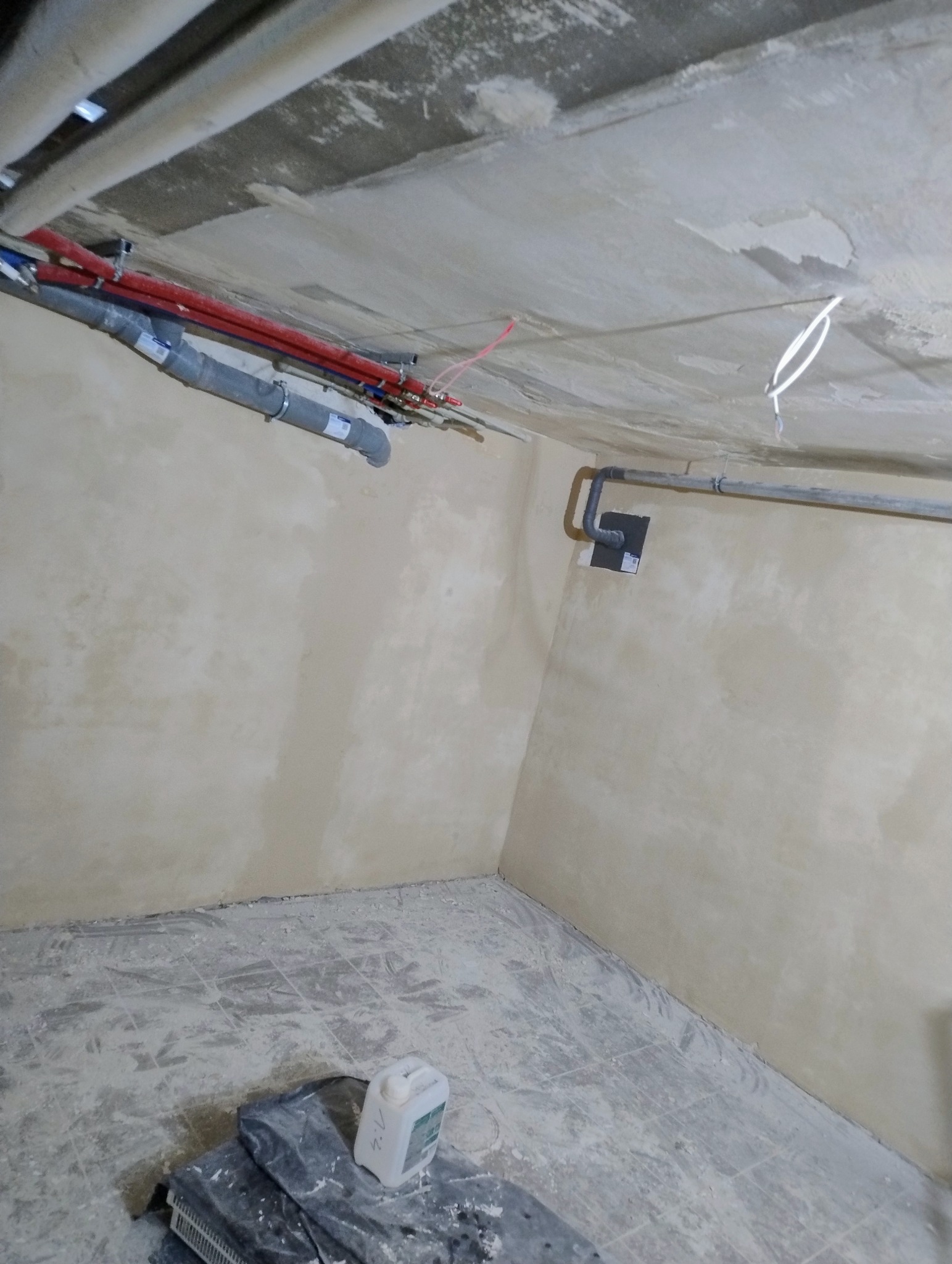 Pokój po szpachlowaniu ścian i sufitu, z widocznymi instalacjami hydraulicznymi pod sufitem. Podłoga zabezpieczona folią, widoczne ślady gładzi.