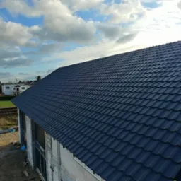 Częściowo pokryty, ciemnogranatowy dach z blachodachówki modułowej, widoczne łaty i membrana, w tle zabudowa jednorodzinna i pochmurne niebo.