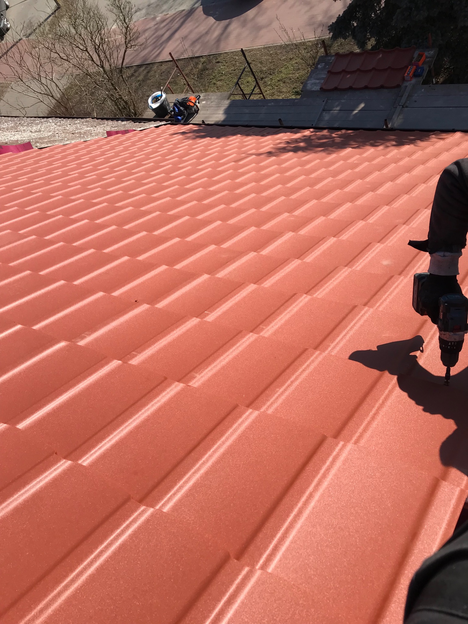 Montaż blachodachówki w kolorze ceglastym, widoczny fragment dachu z narzędziem w trakcie pracy, w tle drzewo bez liści i budynki.