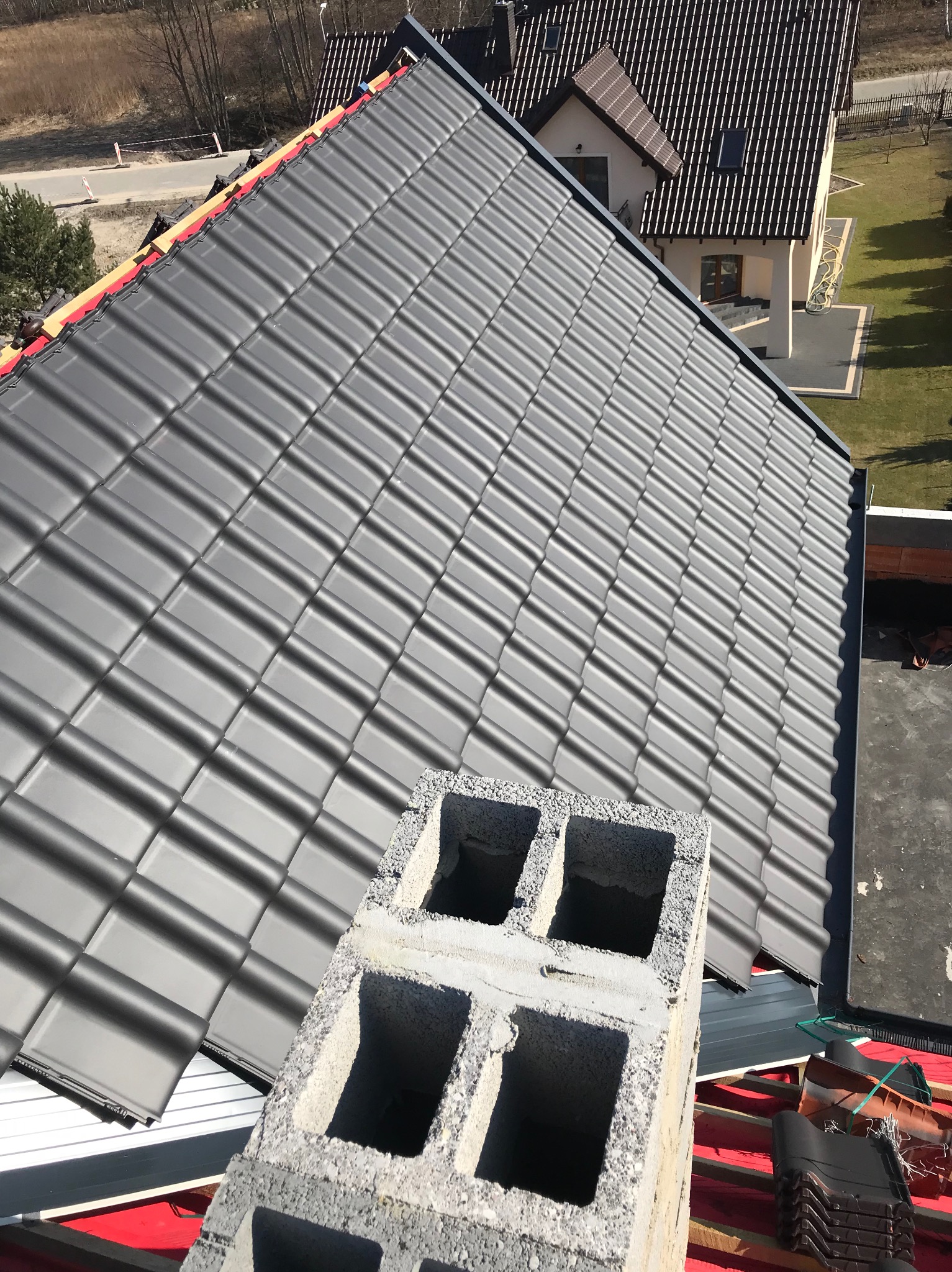 Widok z góry na nowo kryty dach z szarej dachówki ceramicznej, z widocznym kominem w trakcie budowy z pustaków betonowych i fragmentem dachu sąsiedniego domu w tle.