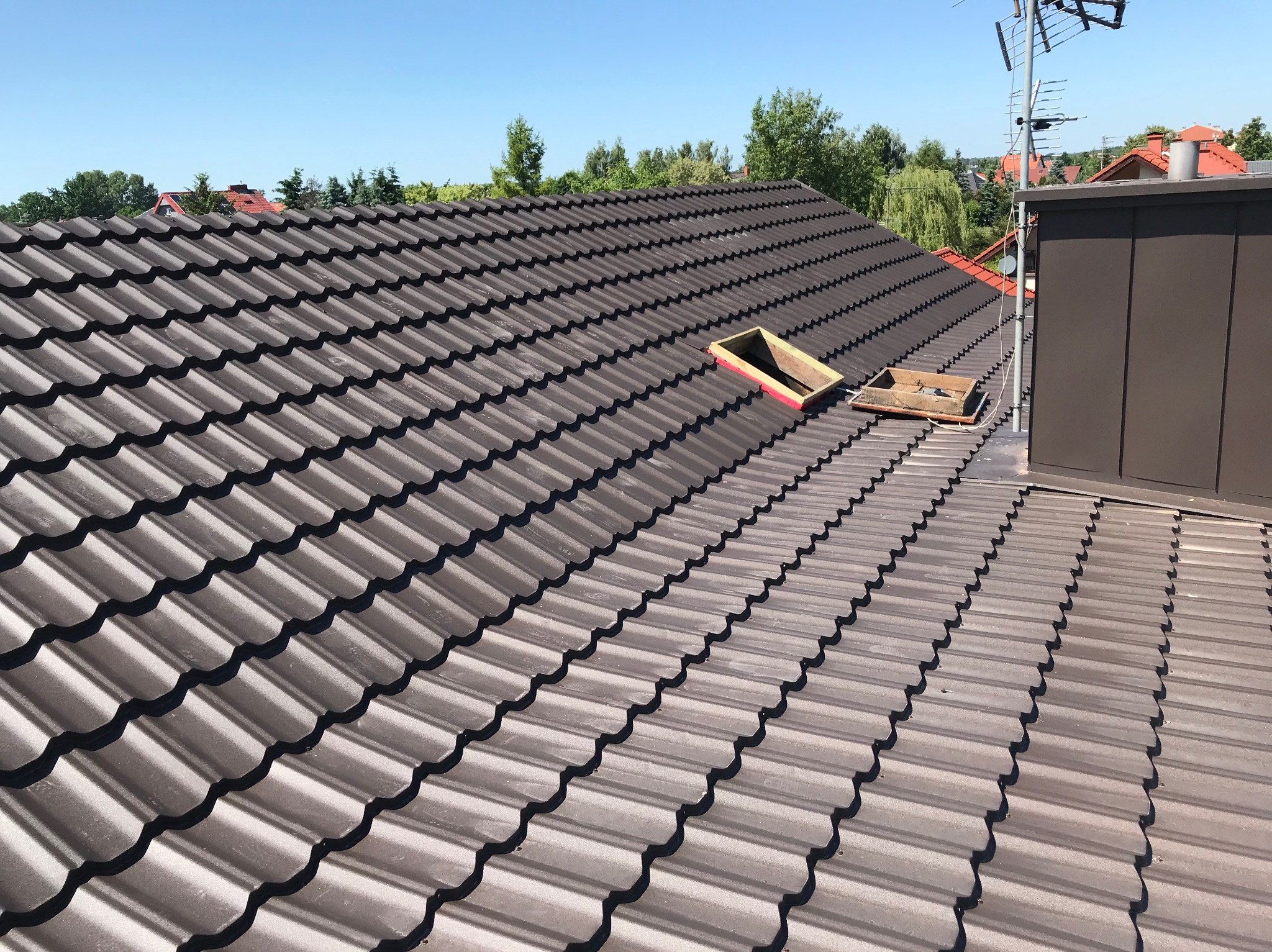 Brązowy dach z dachówki ceramicznej, z widocznymi dwoma otworami okien dachowych w trakcie montażu, antena telewizyjna w tle.