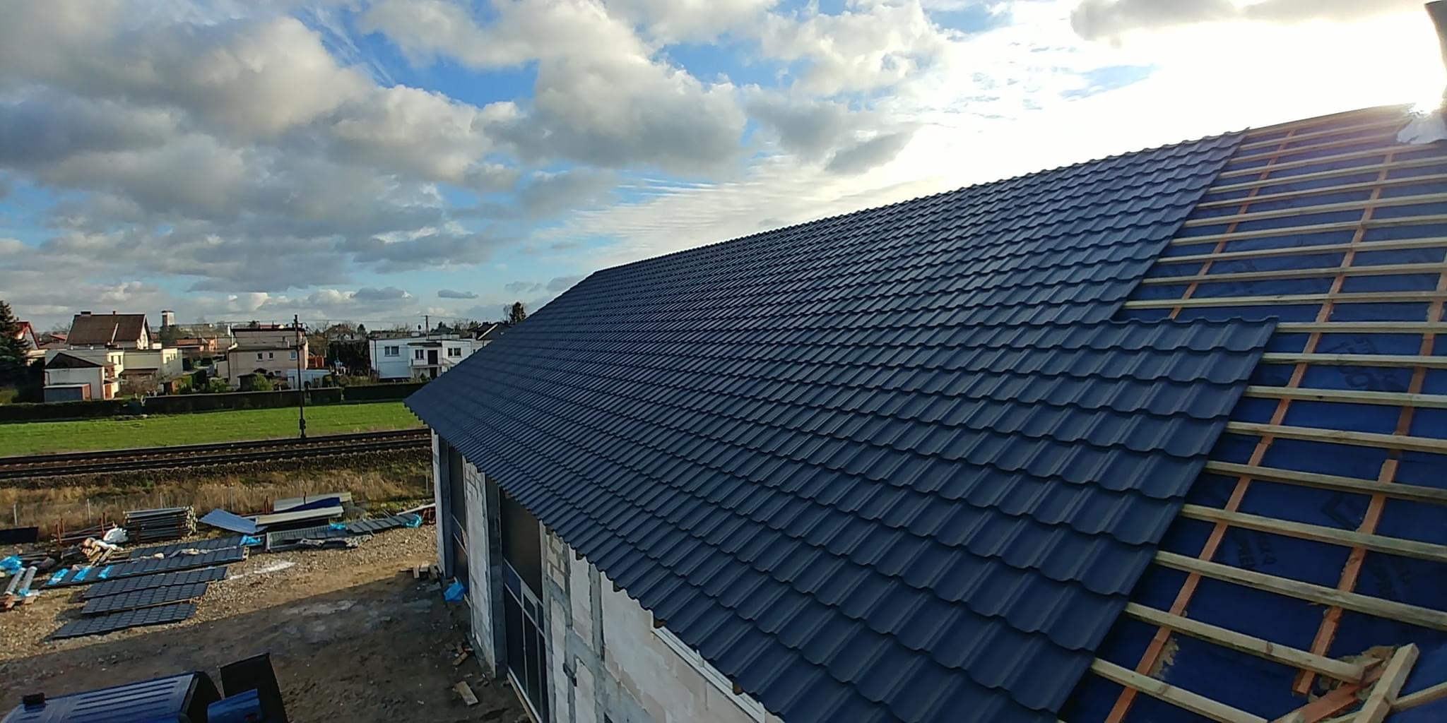 Częściowo pokryty, ciemnogranatowy dach z blachodachówki modułowej, widoczne łaty i membrana, w tle zabudowa jednorodzinna i pochmurne niebo.