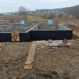 Budowa fundamentów domu z bloczków betonowych, zabezpieczonych czarną izolacją, na błotnistym terenie, z widocznym pracownikiem w tle.