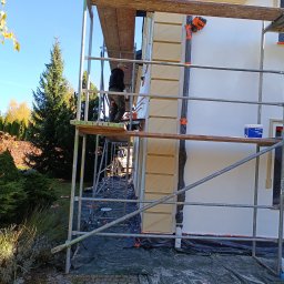 DG Monika Kaleta - Pracownik na rusztowaniu maluje elewację domu z dekoracyjnymi elementami, zabezpieczoną folią, widoczna puszka z farbą, w tle drzewa i błękitne niebo.