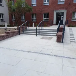 Wejście do budynku z czerwonej cegły z rampą i schodami wyłożonymi szarą kostką, otoczone jasnoszarą kostką brukową i niskim murem z cegły, widoczne gołębie.