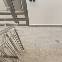 Wnętrze klatki schodowej z metalową balustradą, szarą, przeszkloną ścianą działową z drzwiami, oraz wykończoną szarą płytką podłogą i schodami.