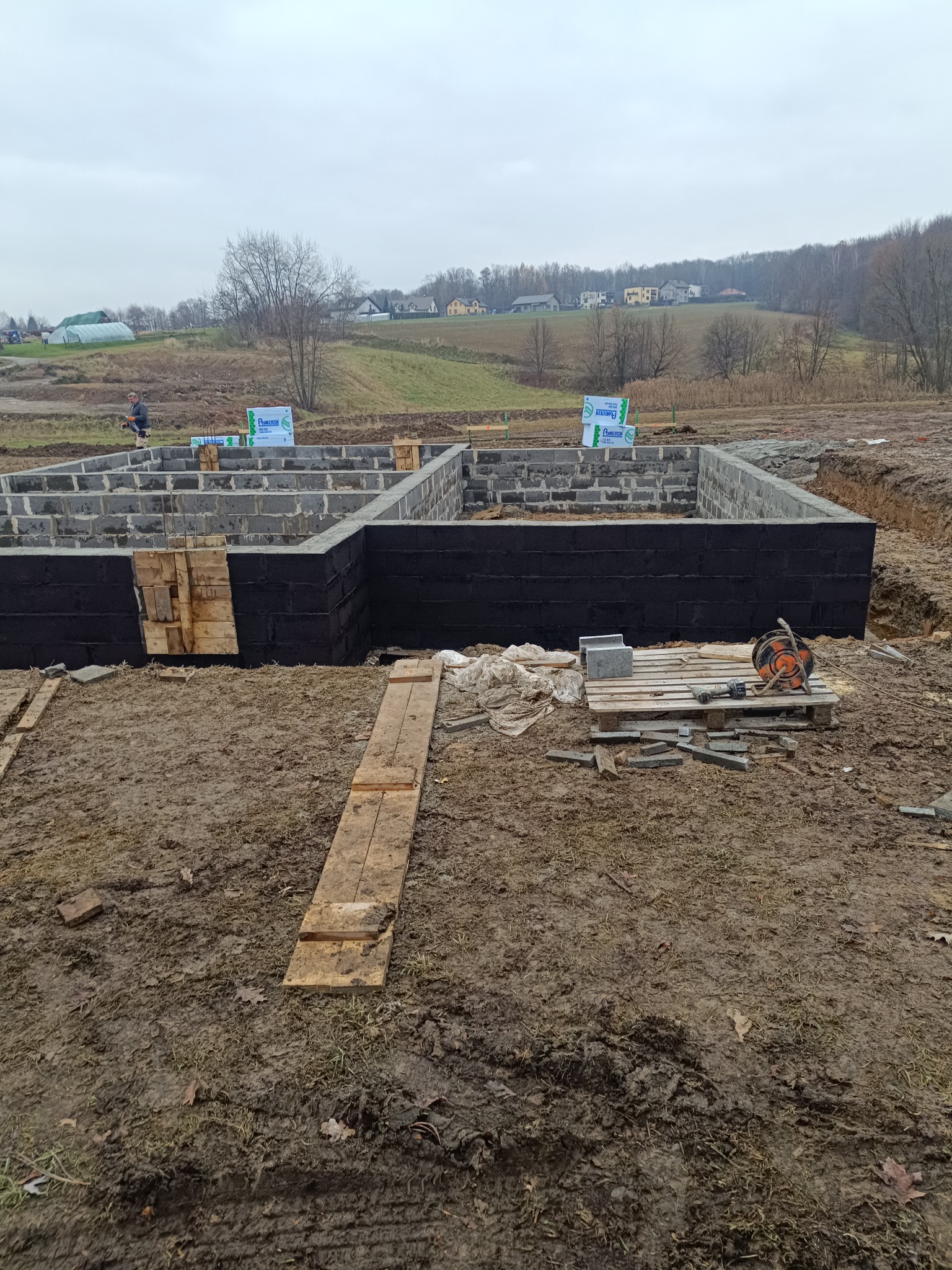 Budowa fundamentów domu z bloczków betonowych, zabezpieczonych czarną izolacją, na błotnistym terenie, z widocznym pracownikiem w tle.