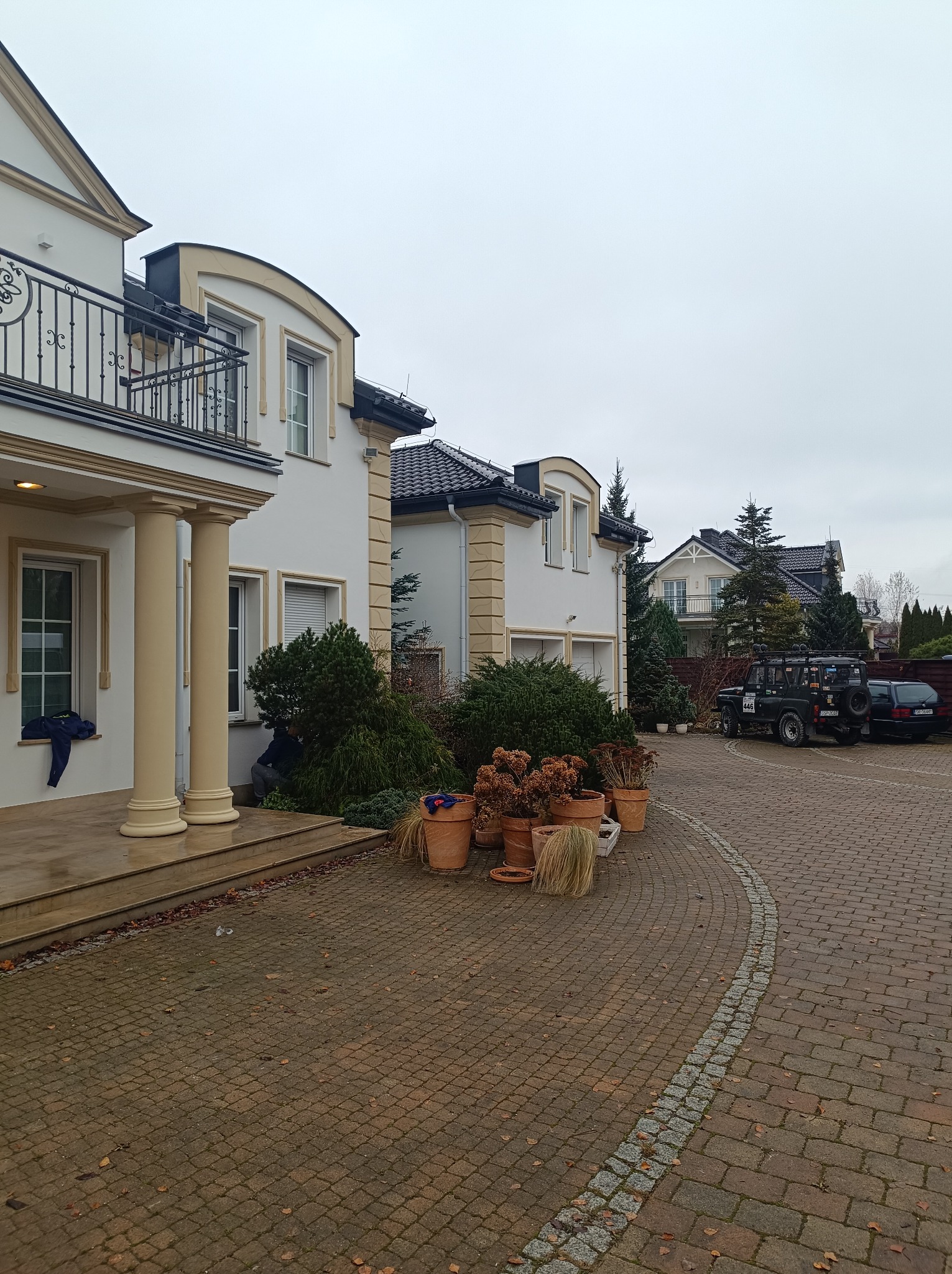 Elegancki dom z kolumnami i balkonem, widok na brukowany podjazd z zaparkowanymi samochodami i dekoracyjnymi donicami, pochmurny dzień.