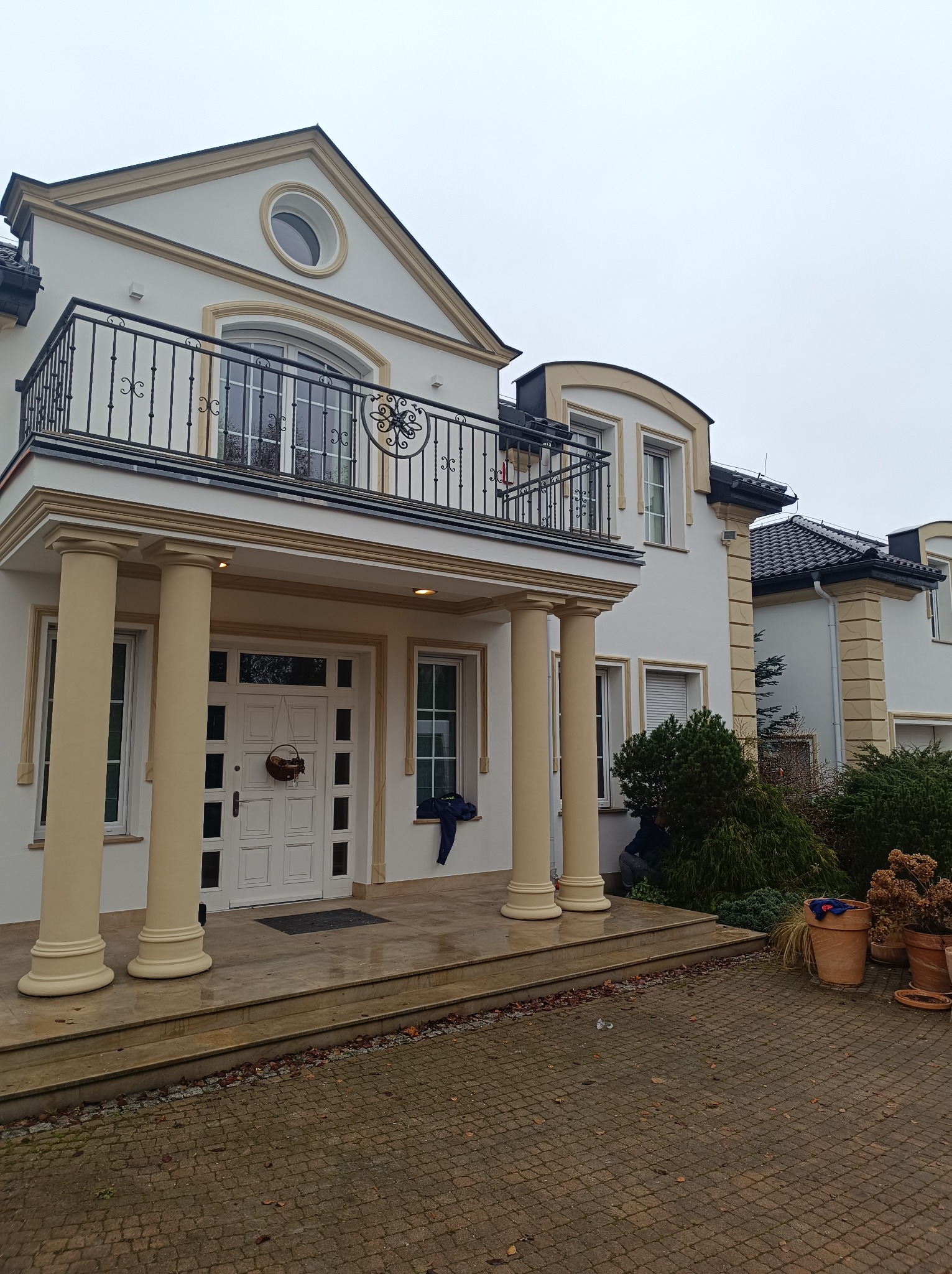 Elegancki, dwupiętrowy dom z kolumnami, balkonem z ozdobną balustradą i okrągłym oknem na szczycie, otoczony brukowanym podjazdem i roślinnością.