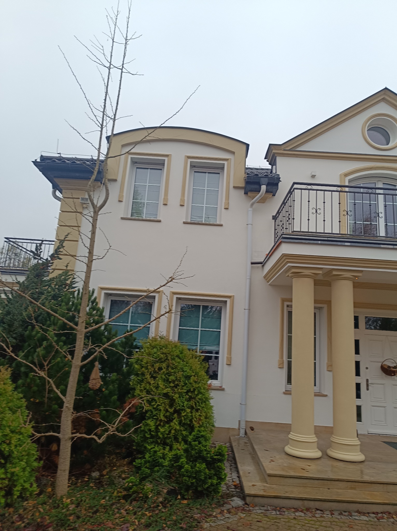Dwupiętrowy dom z jasną elewacją i kolumnami przy wejściu, balkony z ozdobnymi balustradami, widok z poziomu gruntu w pochmurny dzień.