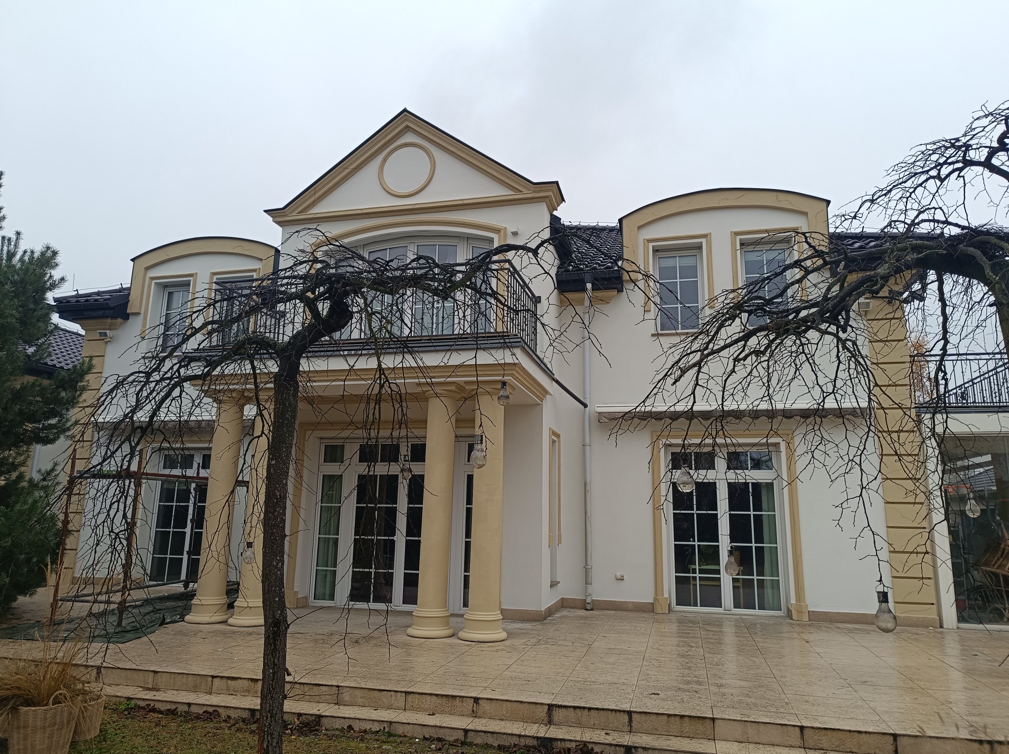Elegancki, dwupiętrowy dom z kolumnami i balkonem, widoczny przez gałęzie drzewa o nagich konarach, z oświetleniem zewnętrznym w postaci wiszących żarówek.