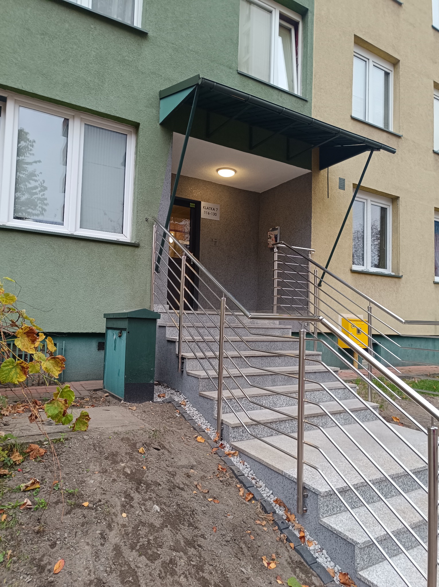 Wejście do budynku wielorodzinnego z nowymi, granitowymi schodami z metalową balustradą, widoczne skrzynki pocztowe i numeracja klatki.