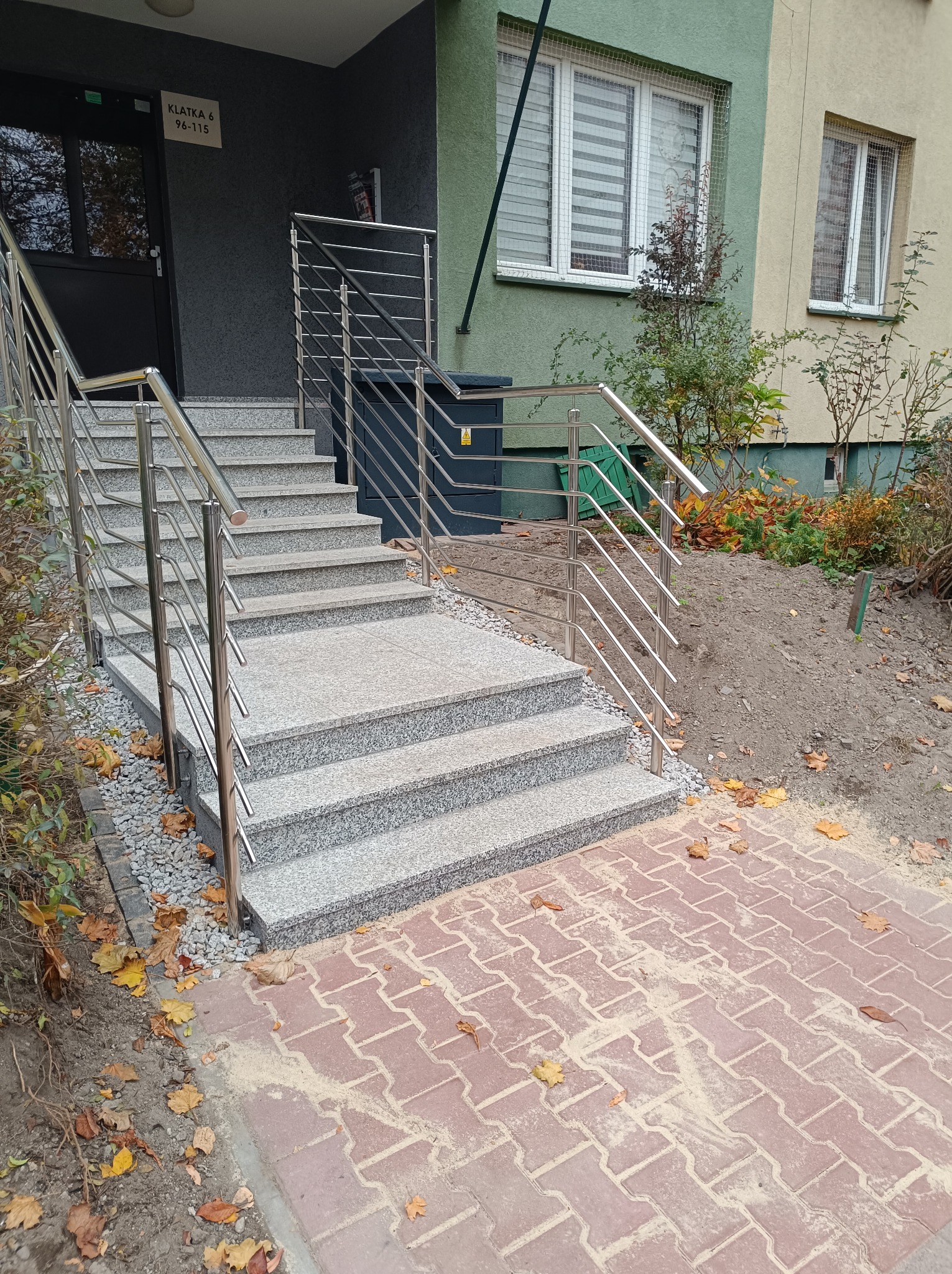 Zewnętrzne schody wejściowe do budynku wielorodzinnego z szarych płyt z metalową balustradą, obok kostka brukowa i opadłe liście.